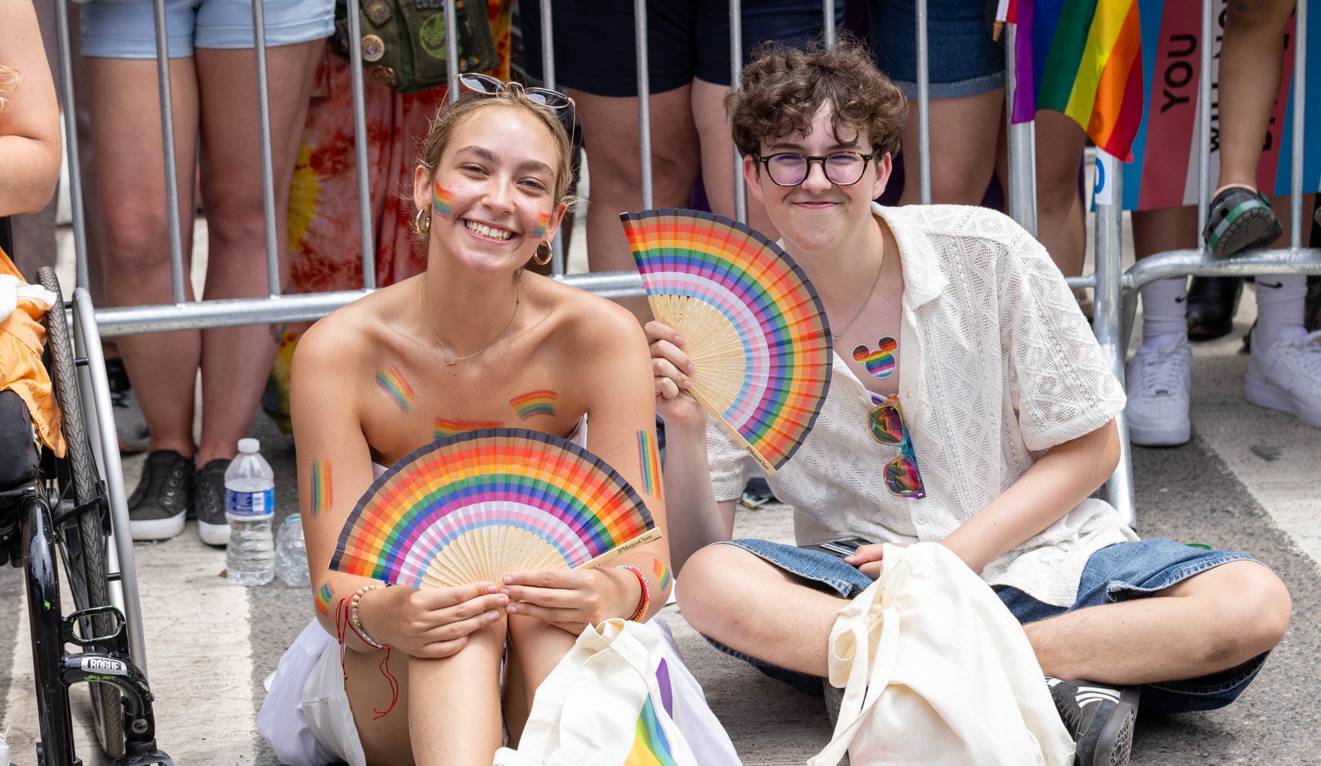 Two smiling people at Pride sit on the street, holding rainbow fans.
