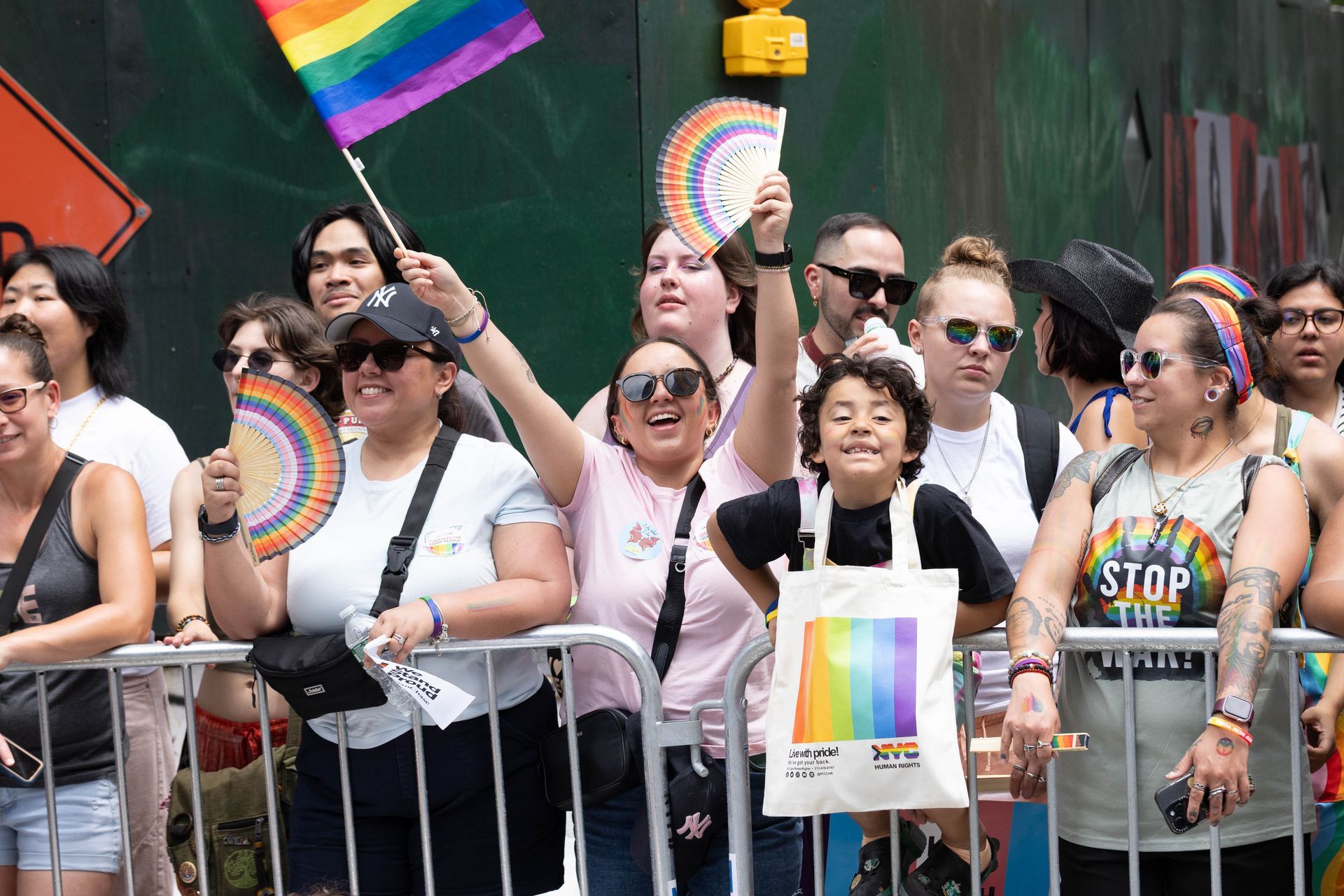 People cheer at a Pride parade, waving rainbow flags and fans behind a metal barrier.