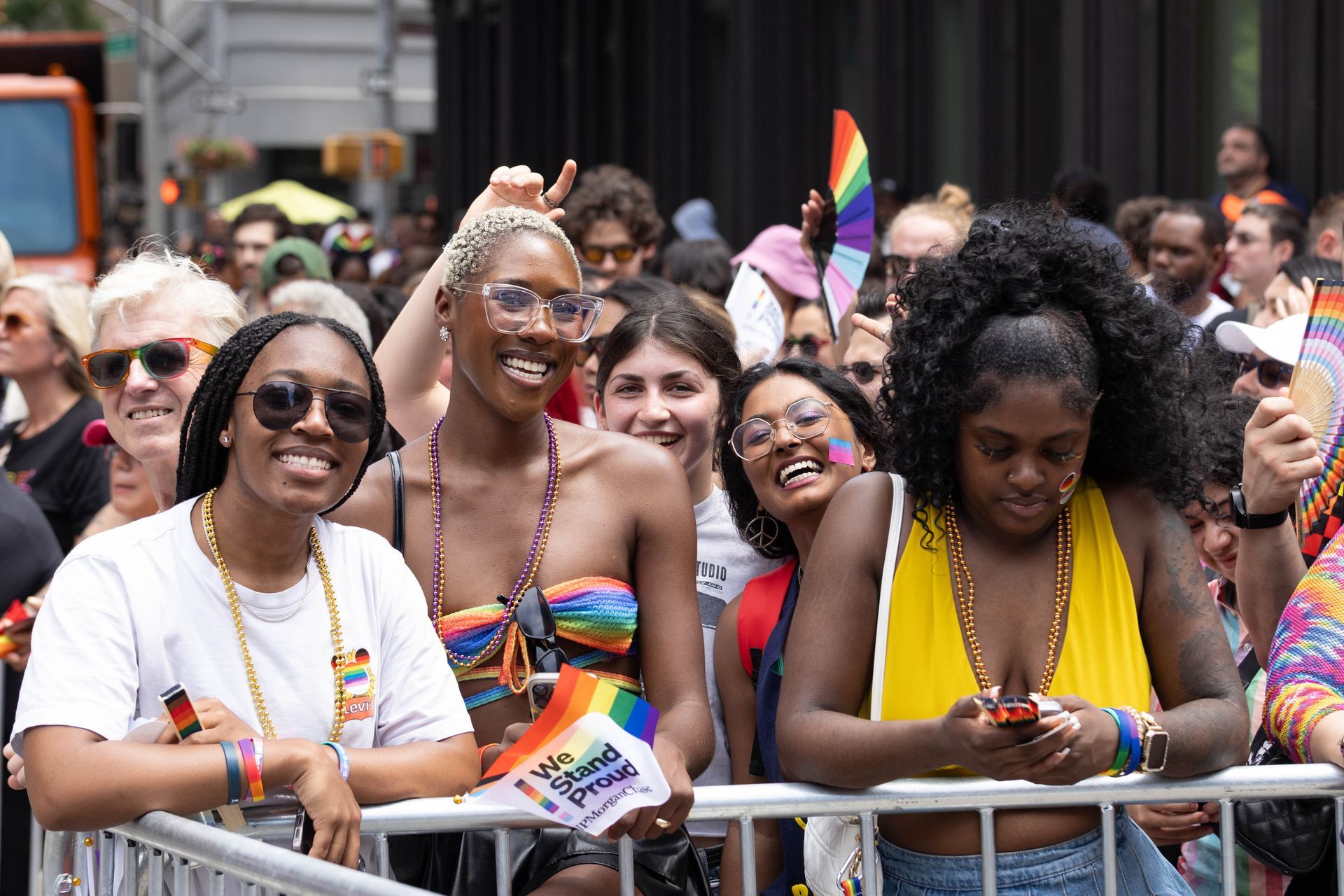 People at a Pride parade, smiling. Rainbow flags & colors, sunny day, crowd behind a barrier.
