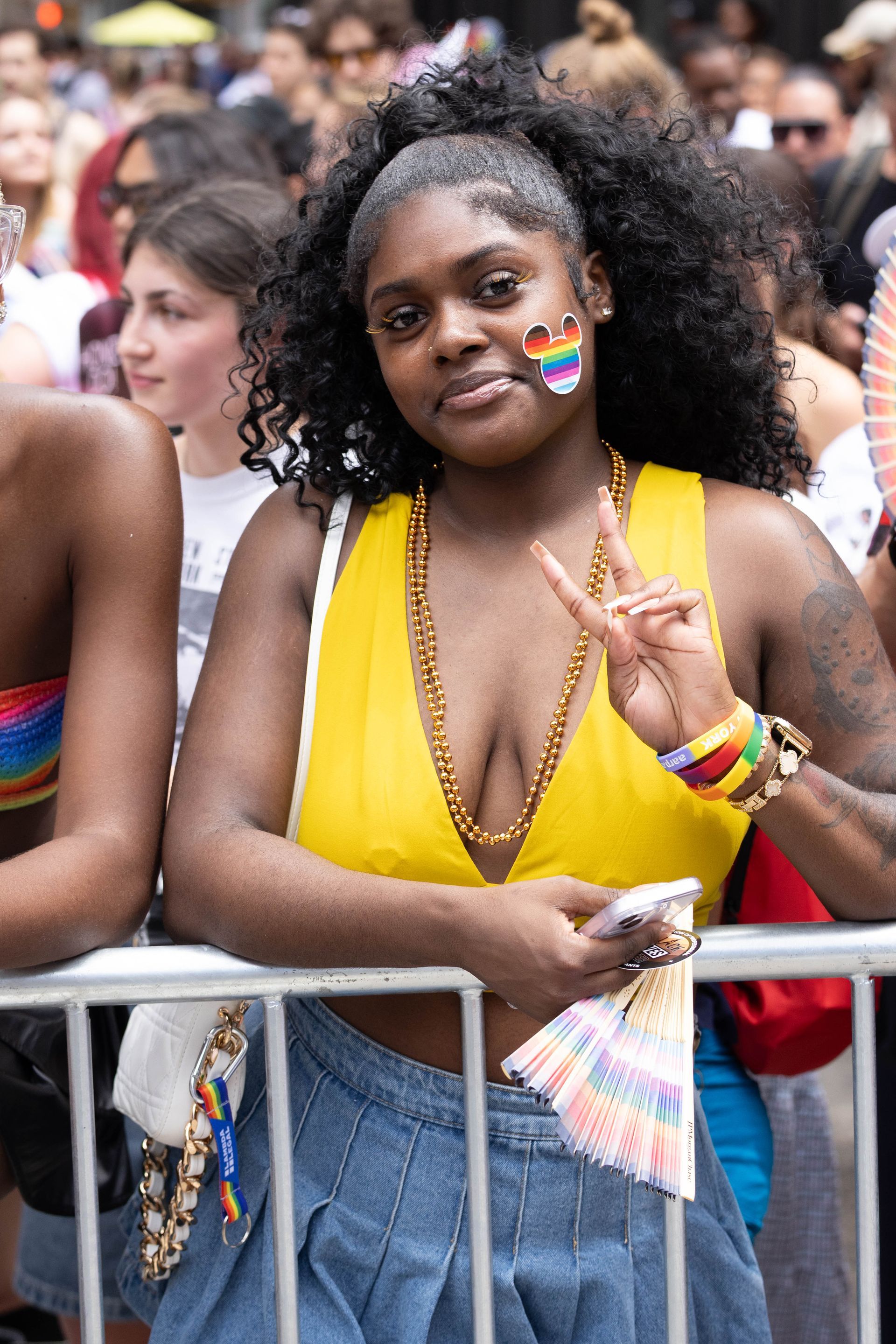 Woman at a pride parade smiles, holding peace sign, with a rainbow flag on cheek and beaded necklace.