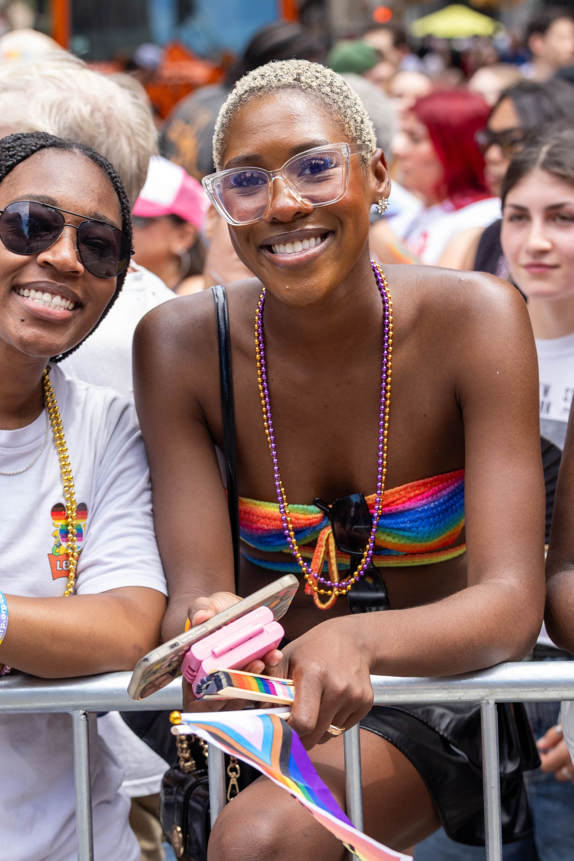 Two smiling Black women at a Pride event, one in a rainbow top, holding a pride flag.