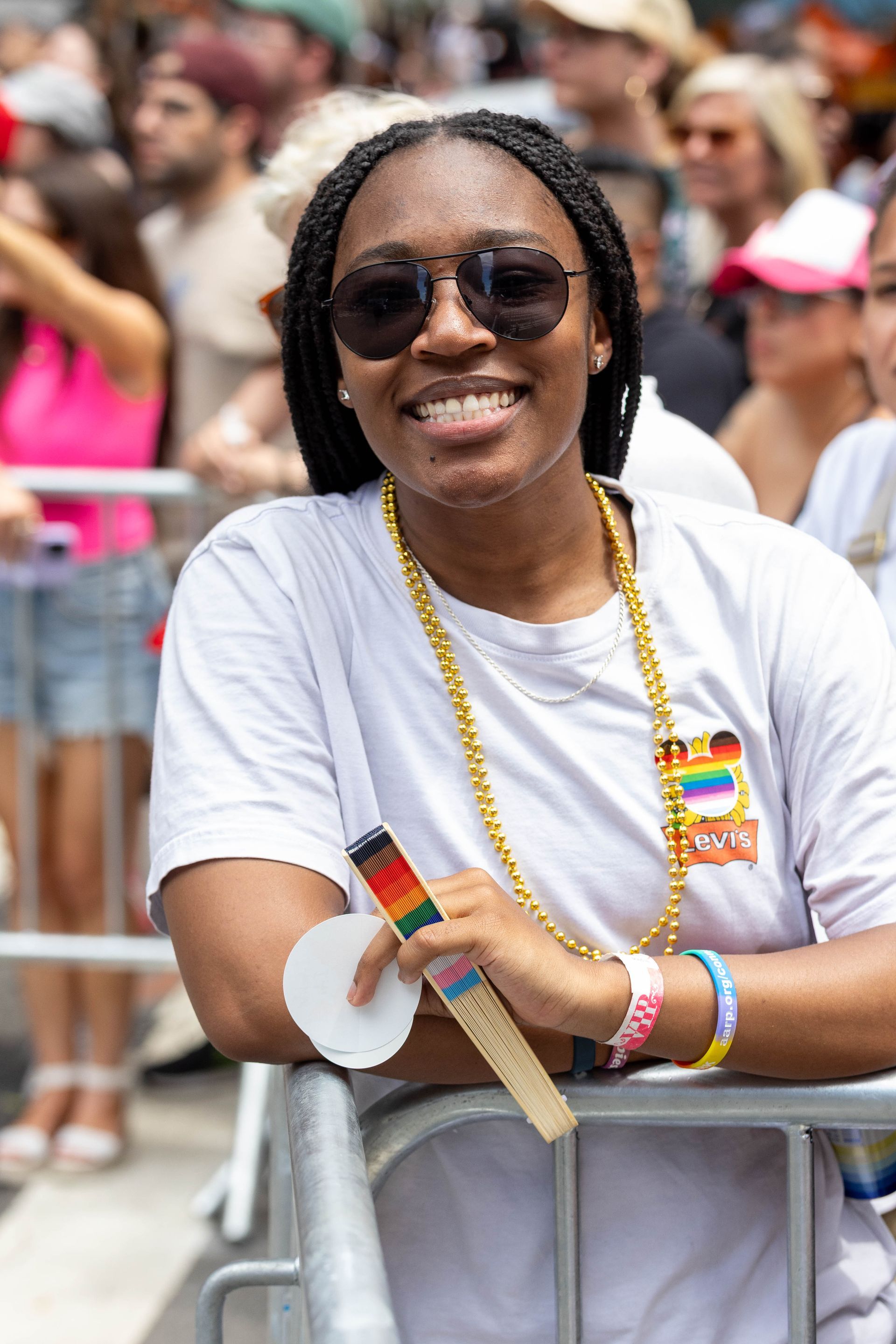Smiling Black woman at a Pride event, wearing sunglasses, gold beads, and a rainbow fan.