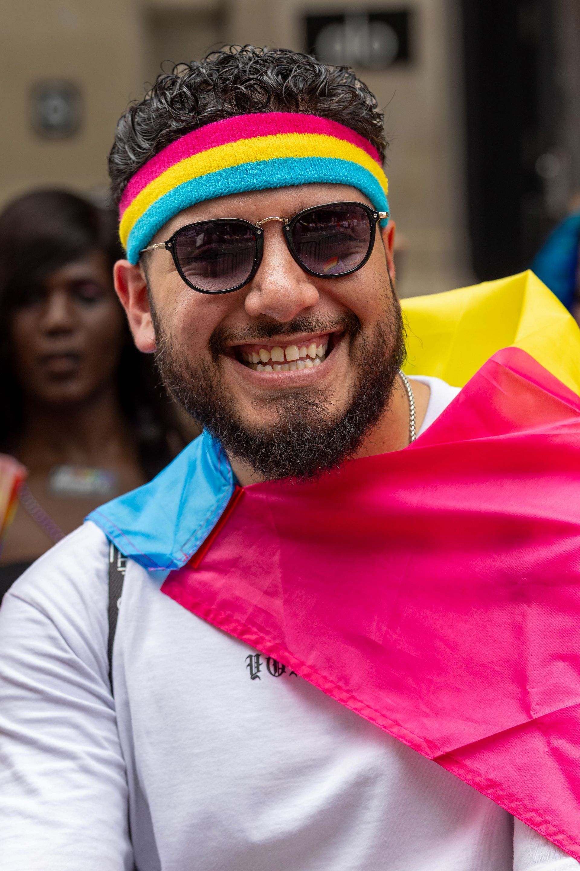 Man smiling, wearing a pansexual flag headband and draped flag, sunglasses.