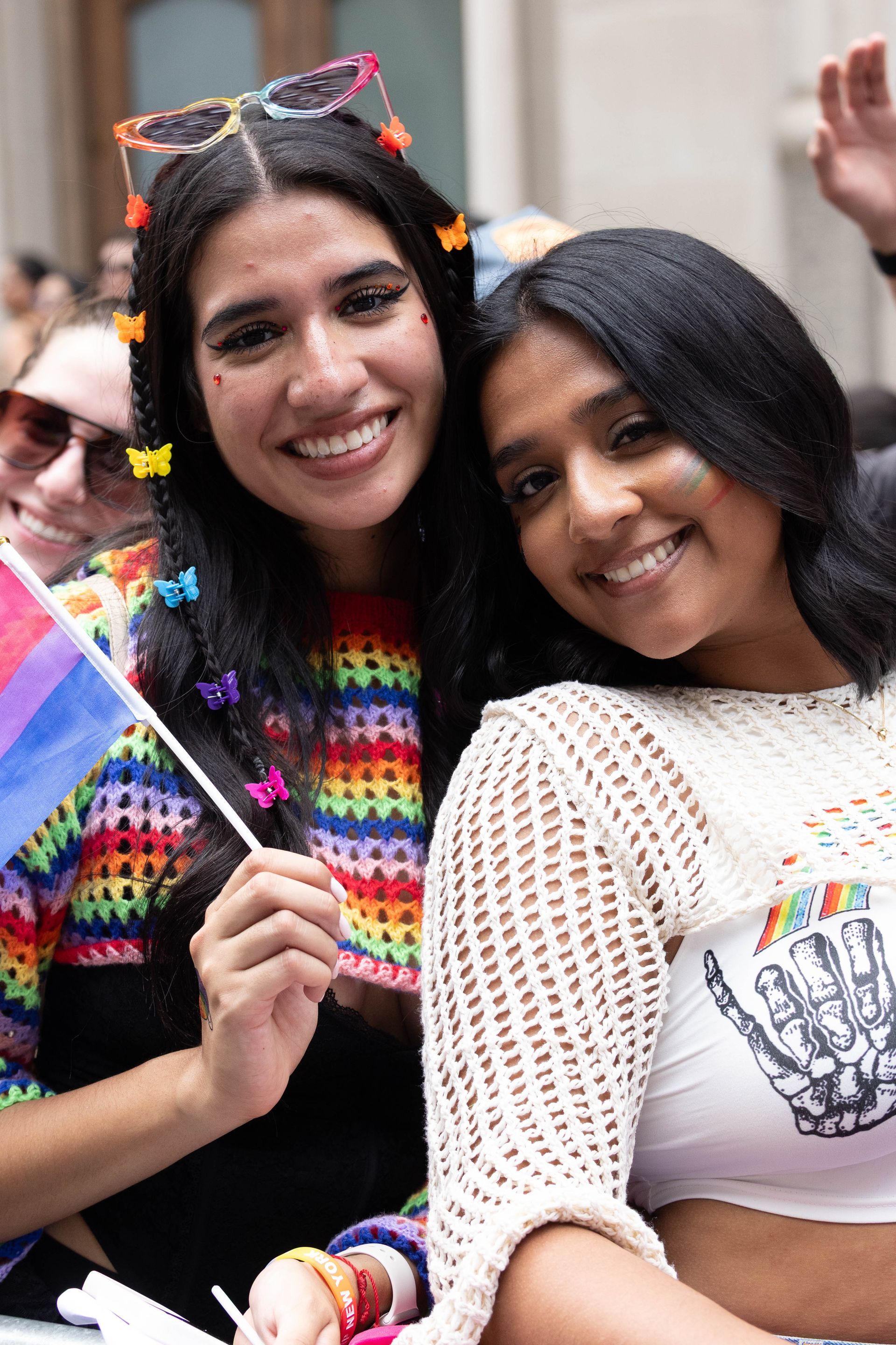 Two smiling women at a pride parade. One holds a bisexual flag, wearing a rainbow top and butterfly clips. The other wears a crocheted top.