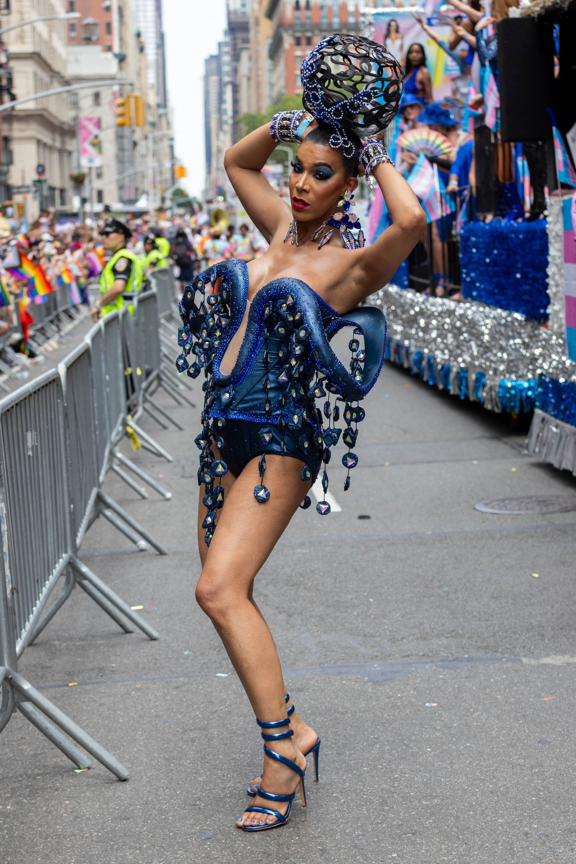 Person in a blue jeweled outfit poses on a float at a parade, New York City.
