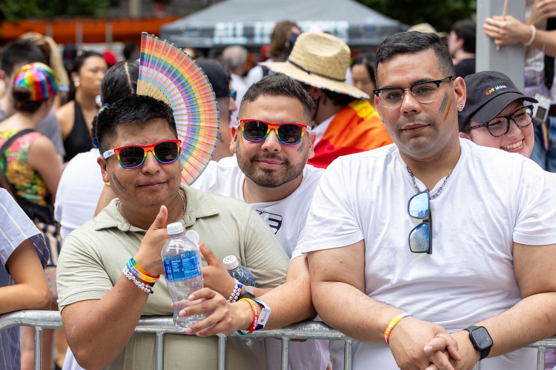 Three men at Pride: two wear rainbow sunglasses, one holds water, all smile.