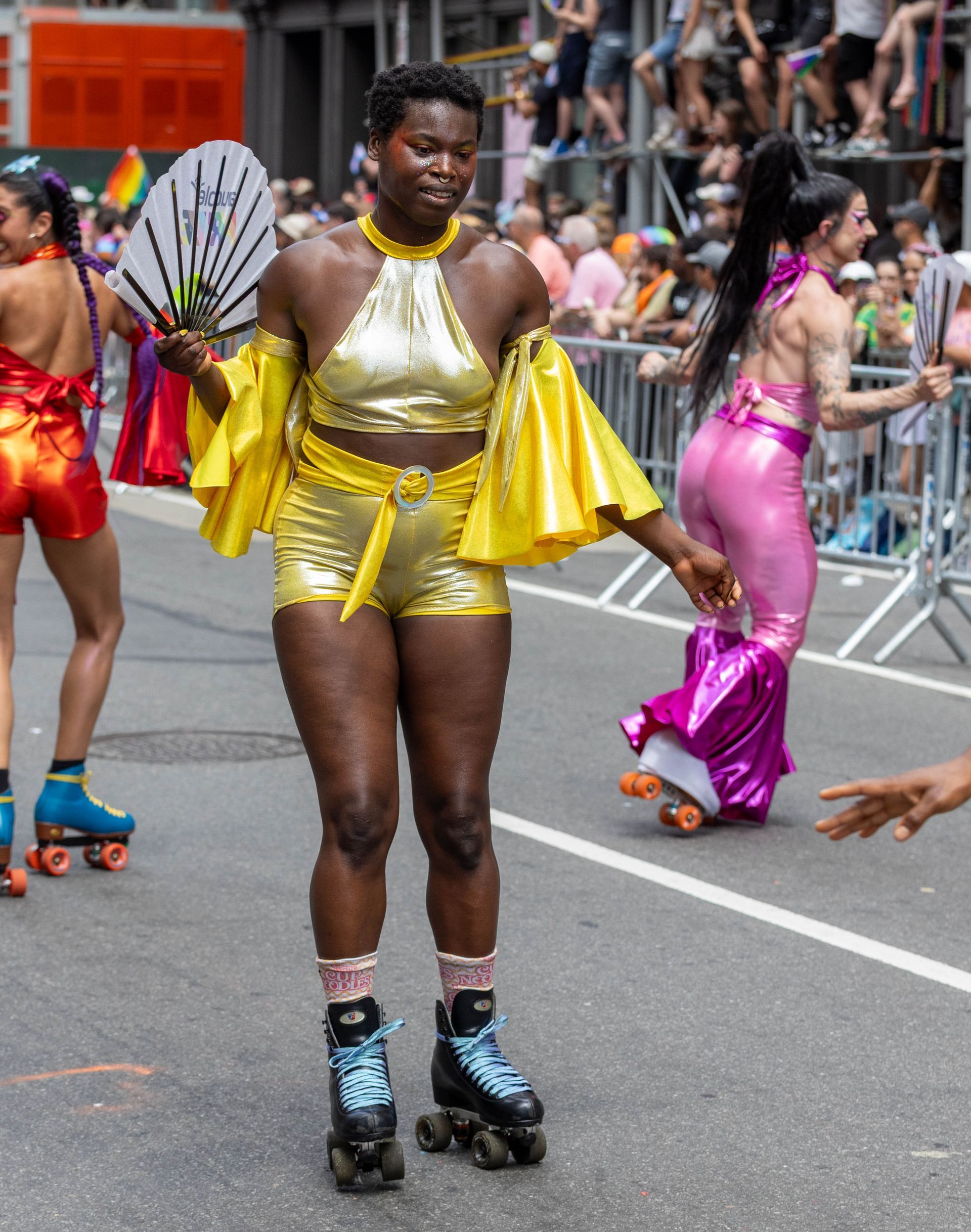 Person in gold outfit on roller skates at Pride parade, holding a fan.