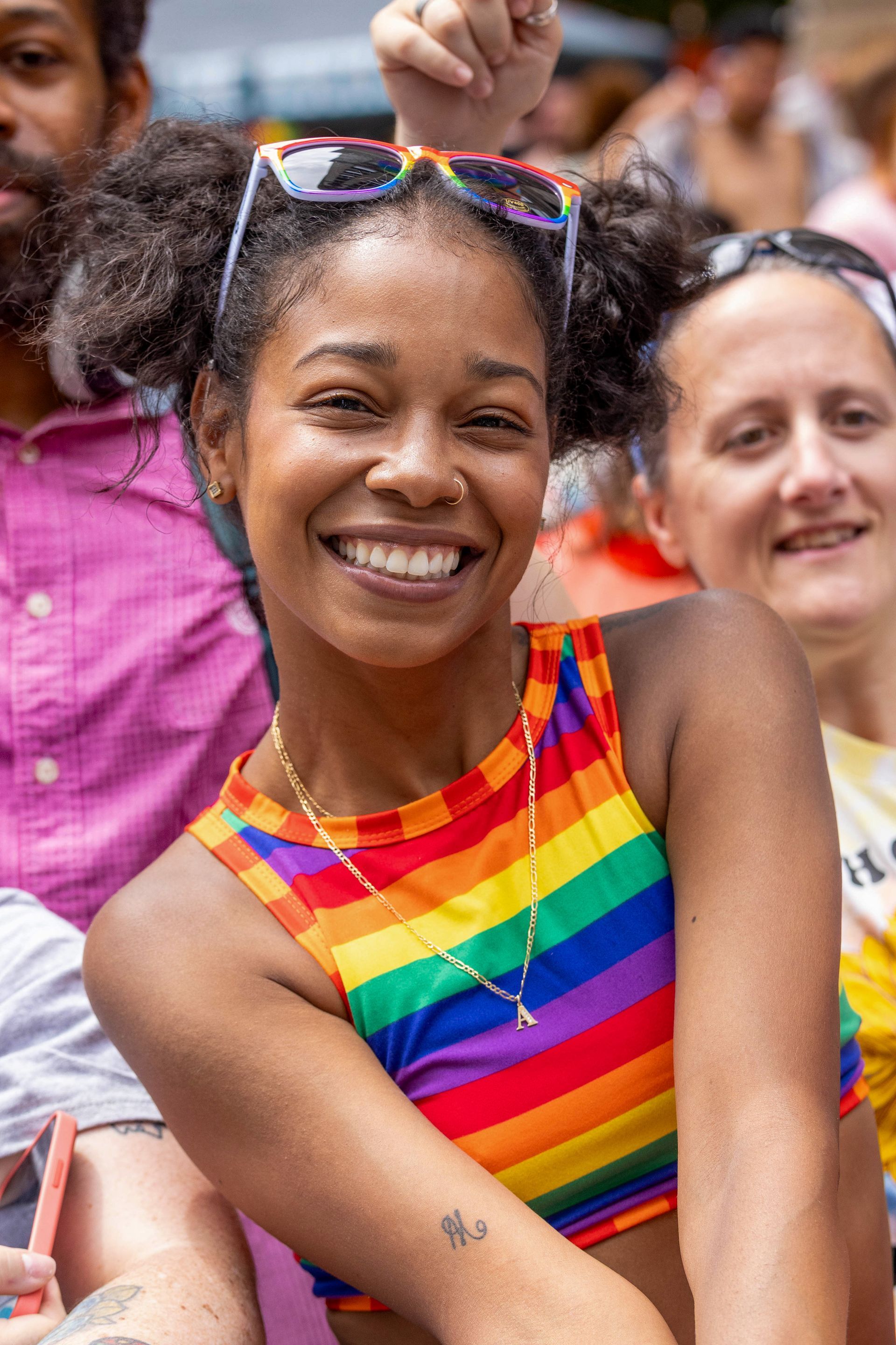 Smiling woman at a Pride event wears rainbow top, sunglasses on head.