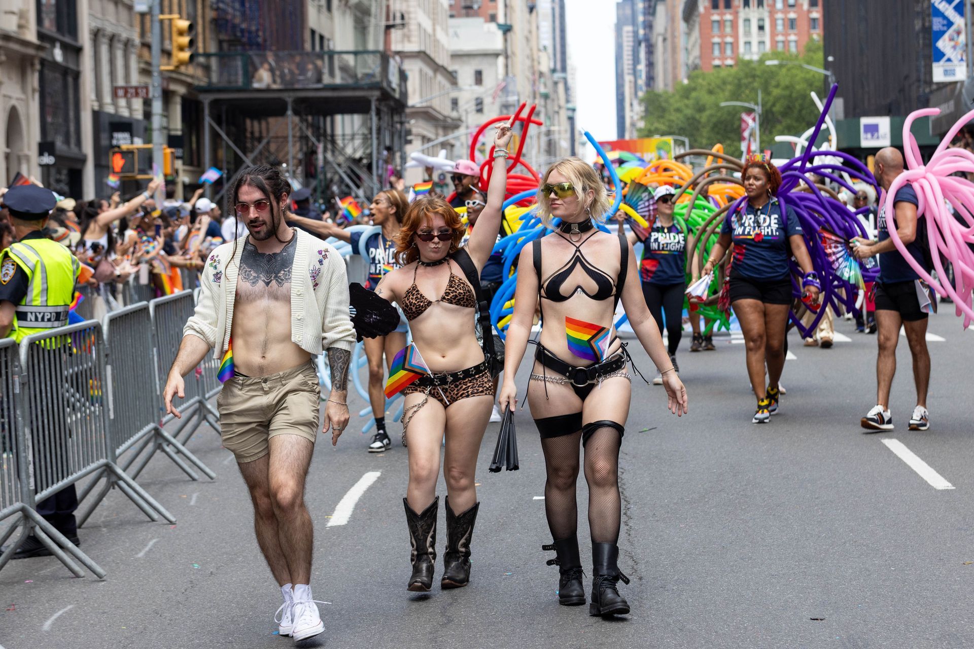 People in flamboyant outfits, celebrating Pride in a city street, smiling and holding rainbow flags.