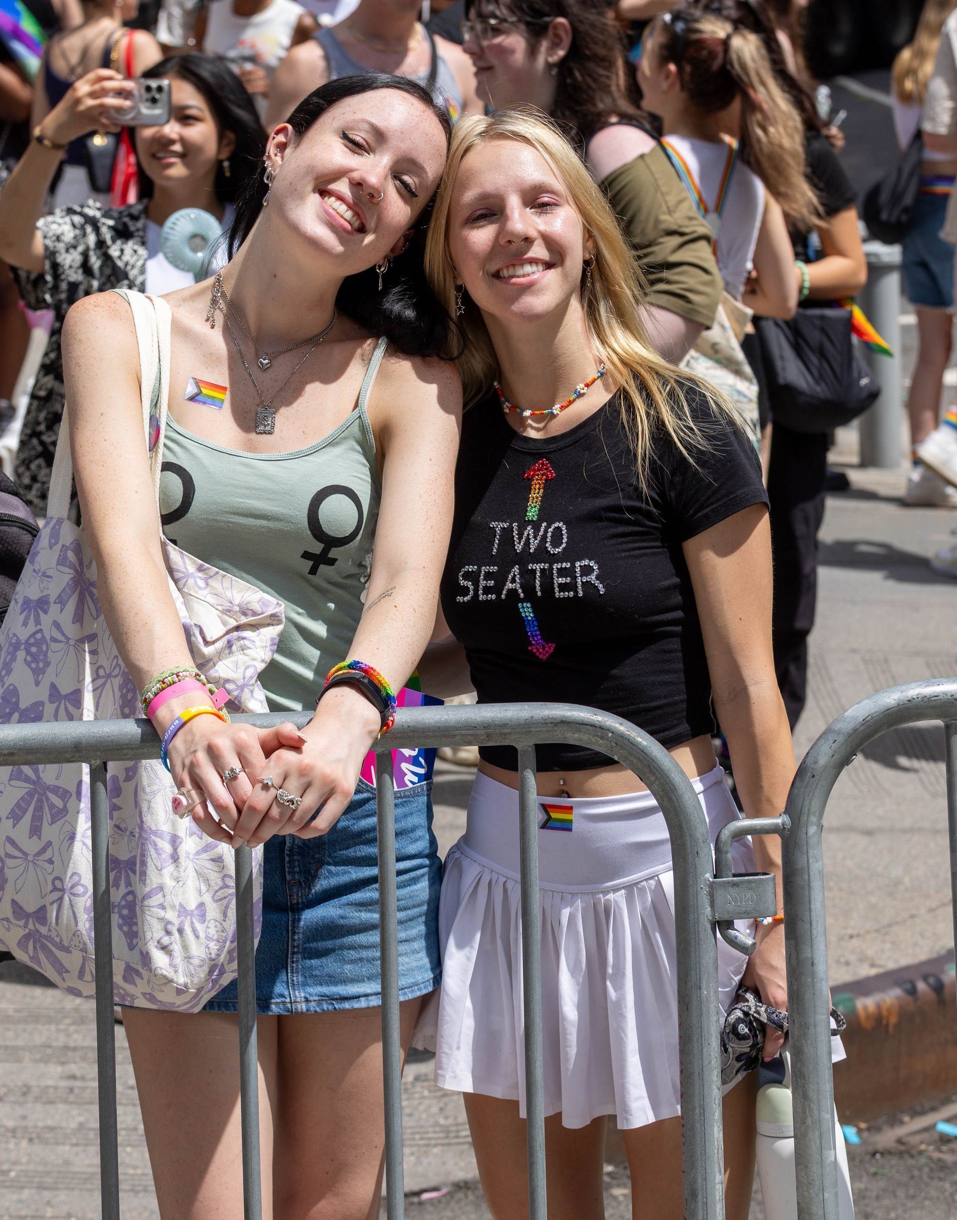 Two smiling young women at a pride event, leaning on a metal barrier; one wears a gender symbol top.