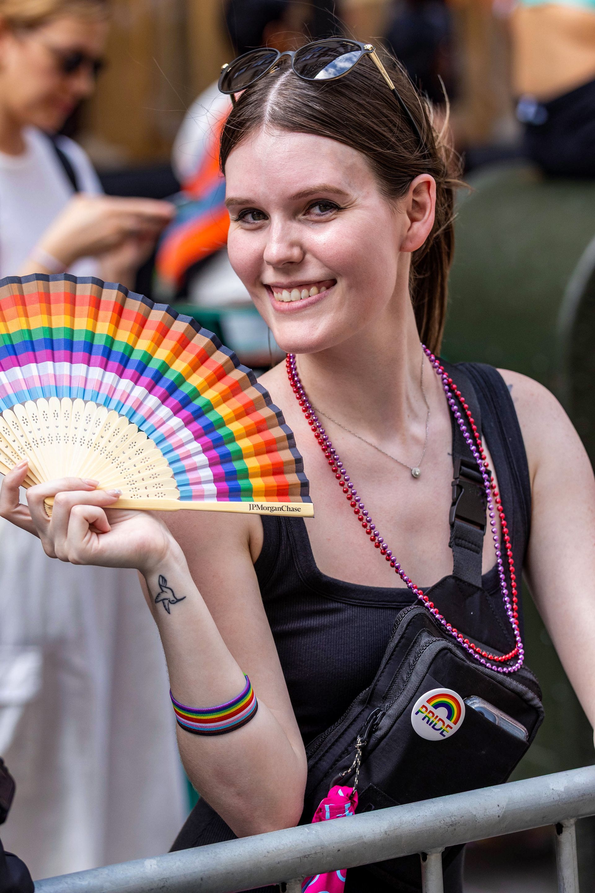 Woman at Pride, holding a rainbow fan, smiling. She has a fanny pack, wristbands, and beads.