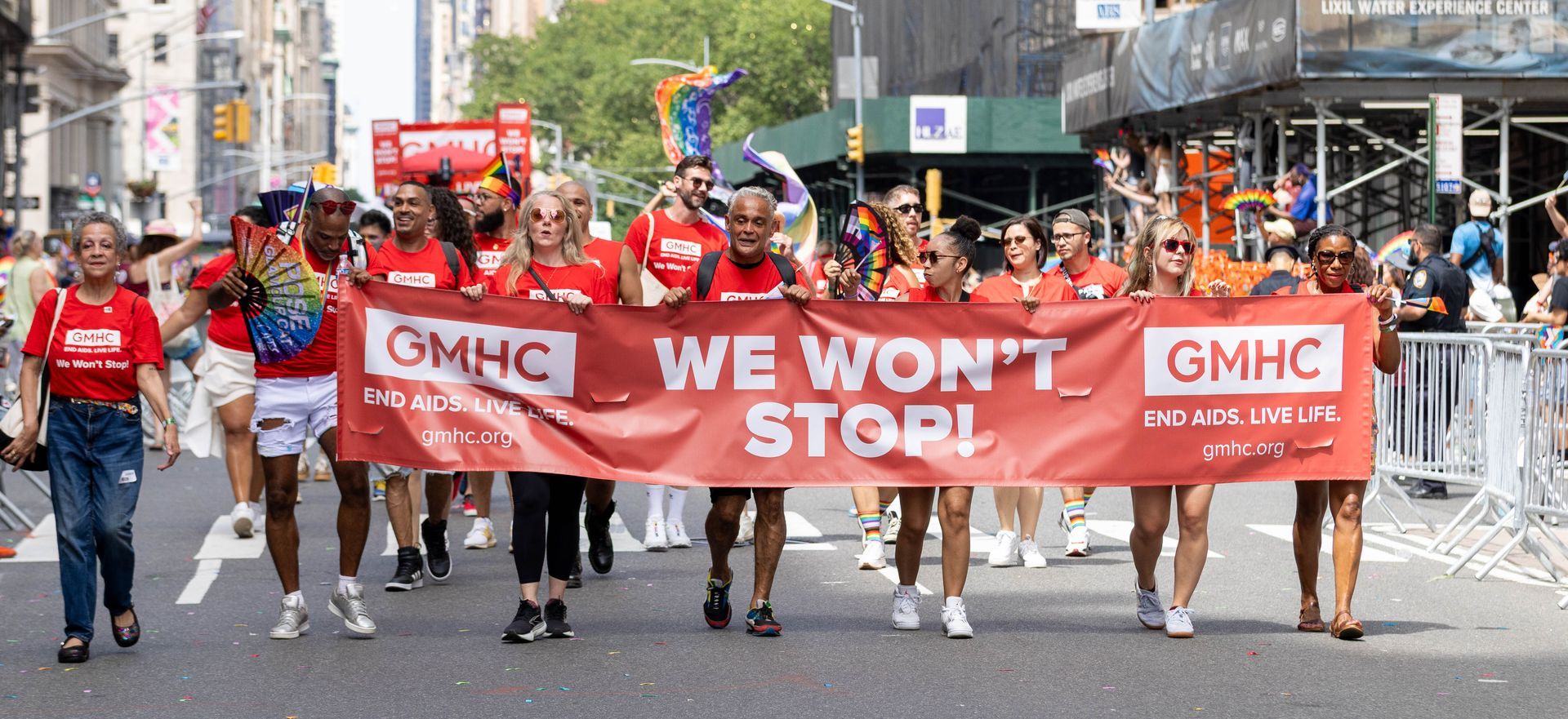People march in a parade, holding a banner that says “WE WON’T STOP!”