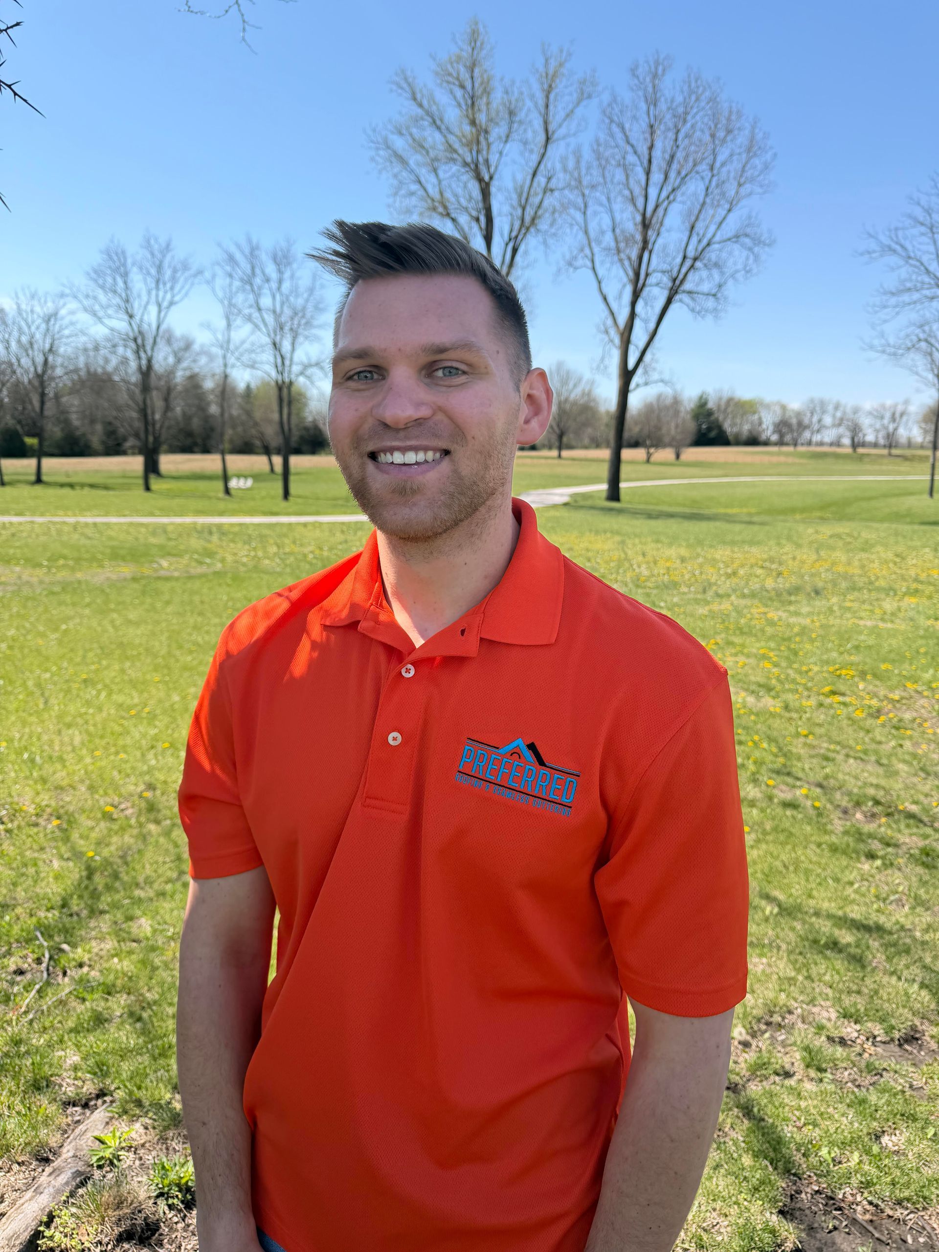 Man in an orange polo shirt smiling in a sunny grassy park