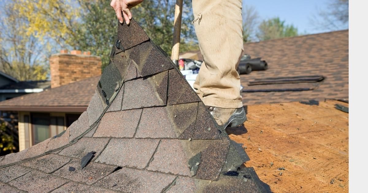 Person on a roof removing asphalt shingles from the ridge. Brown, weathered shingles with a brick chimney in the background.