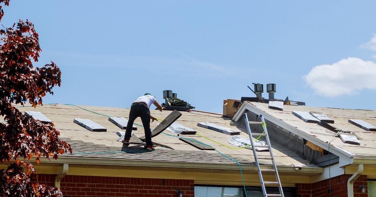 Roofer replacing shingles on a house roof on a sunny day. Ladder and ventilation pipes are visible.