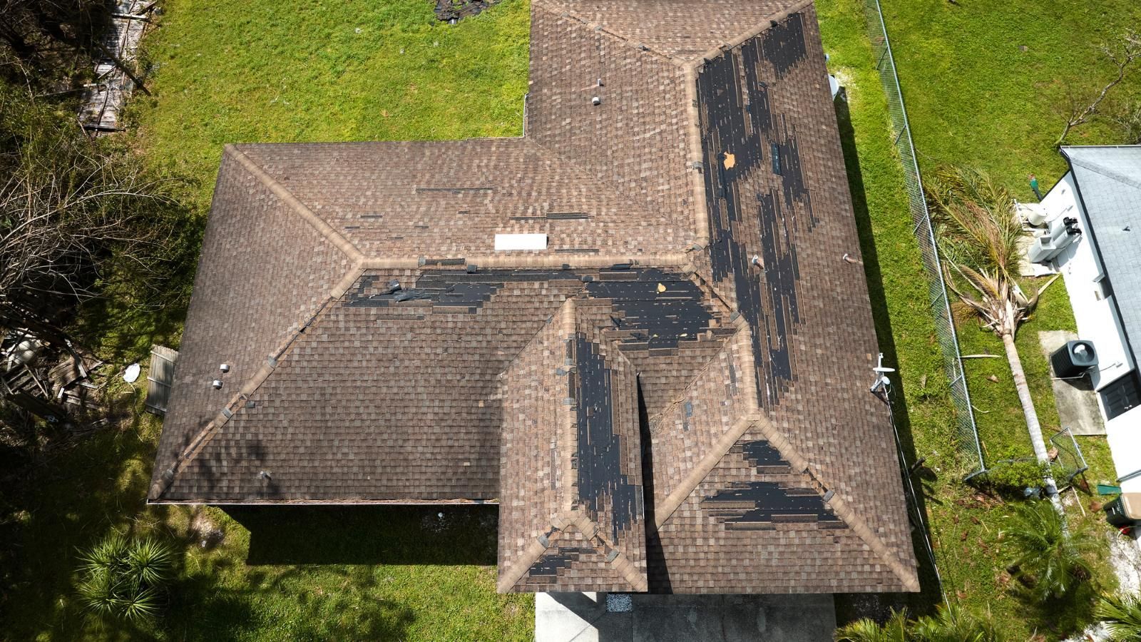 Aerial view of a brown roof with significant damage and missing sections, surrounded by green grass.