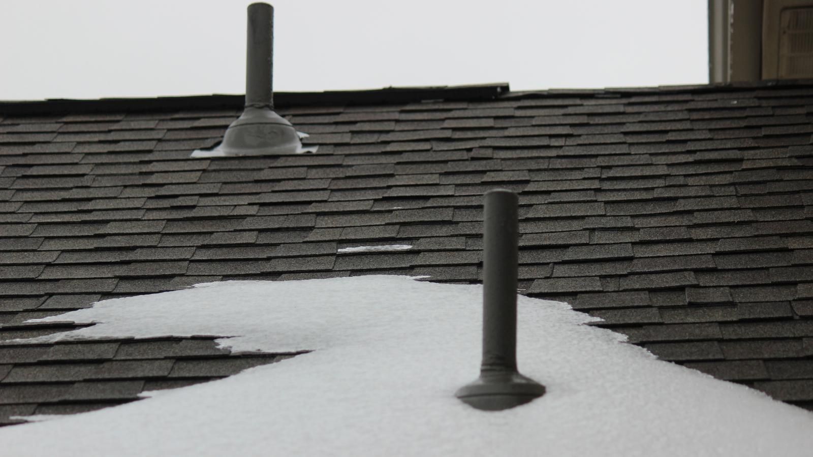 Snow-covered asphalt shingle roof with two vent pipes extending upwards against a cloudy sky.