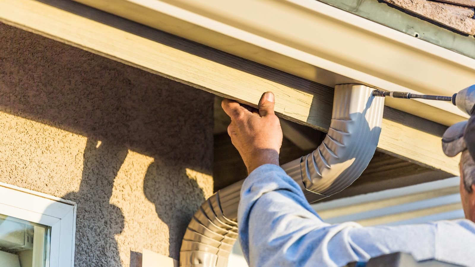Person installs a beige gutter, using a power drill, on a beige house exterior.