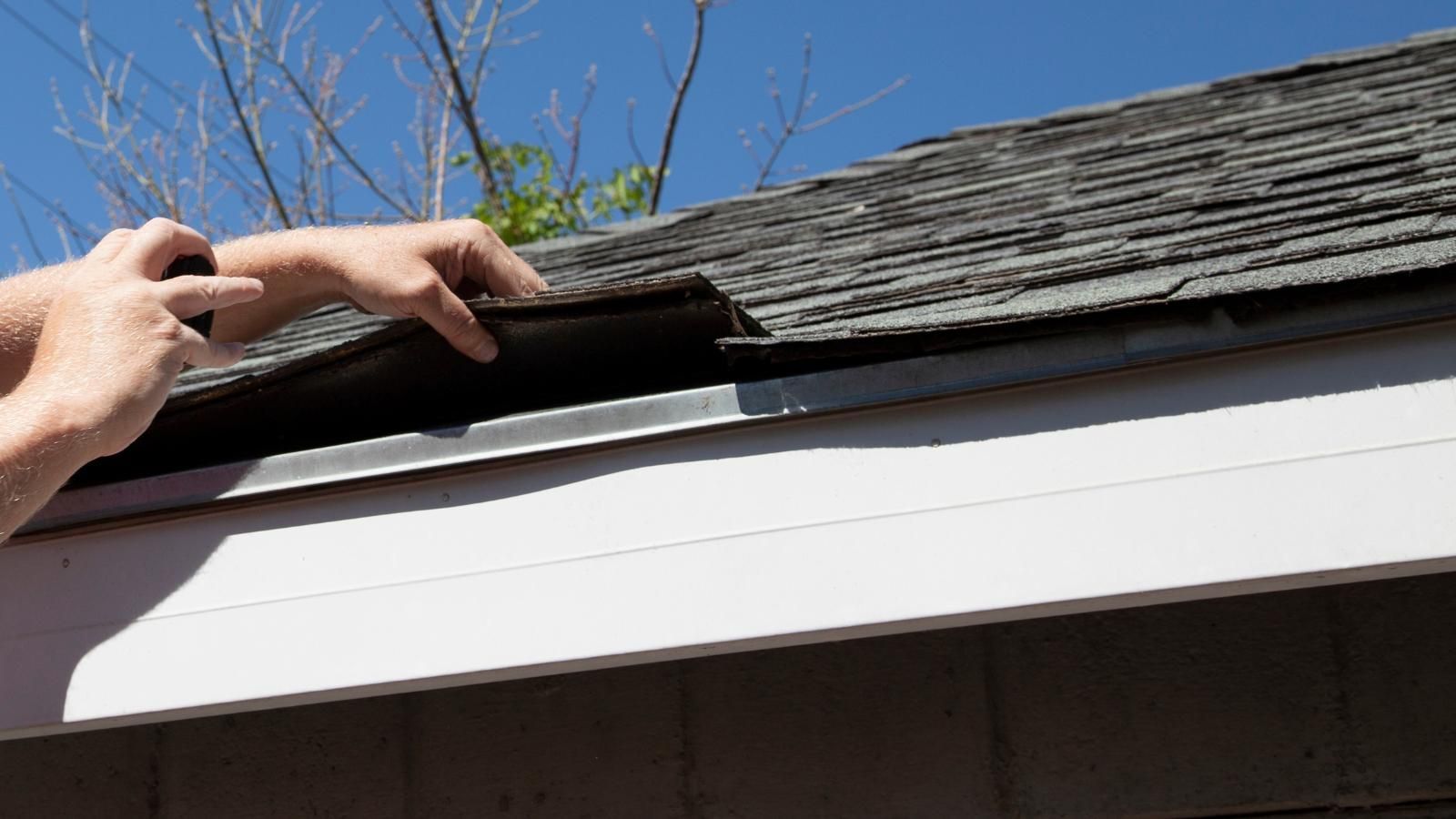 Hands inspecting a roof edge with exposed underlayment under dark shingles