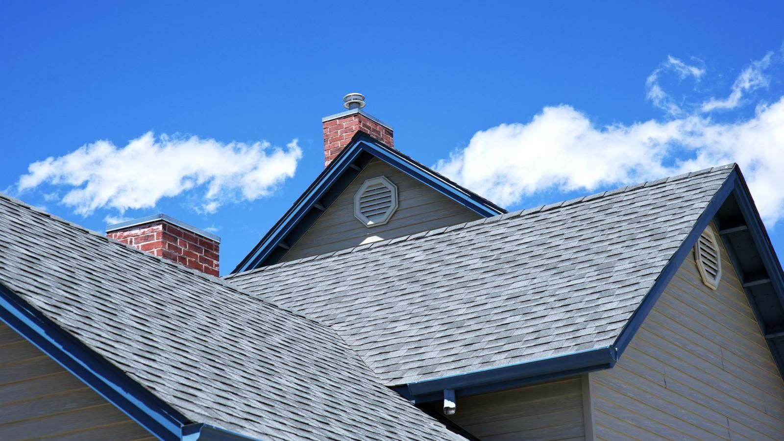Blue-gray shingled roof with dormer and brick chimney against a bright blue sky and clouds