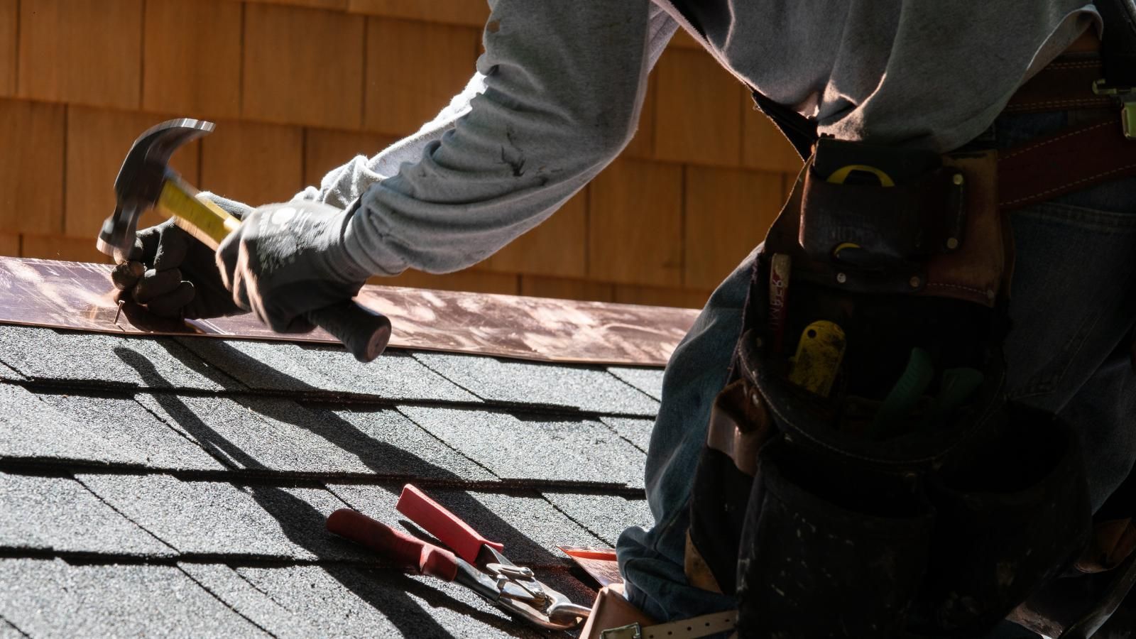 Roofing worker hammering shingles with tools on a sunlit roof