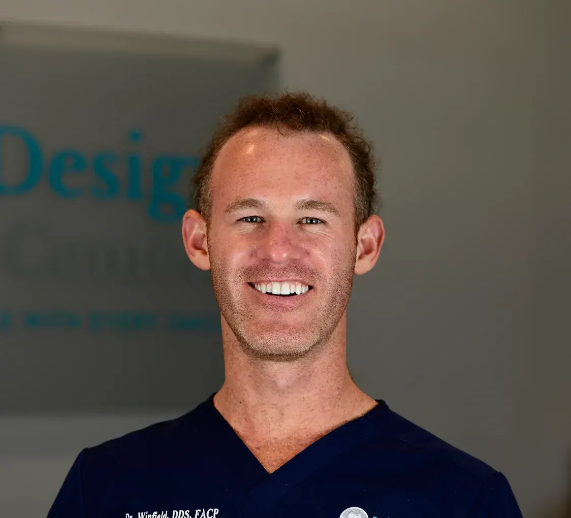 Woman with blonde hair wearing a black turtleneck speaks. Background includes costumed figures.