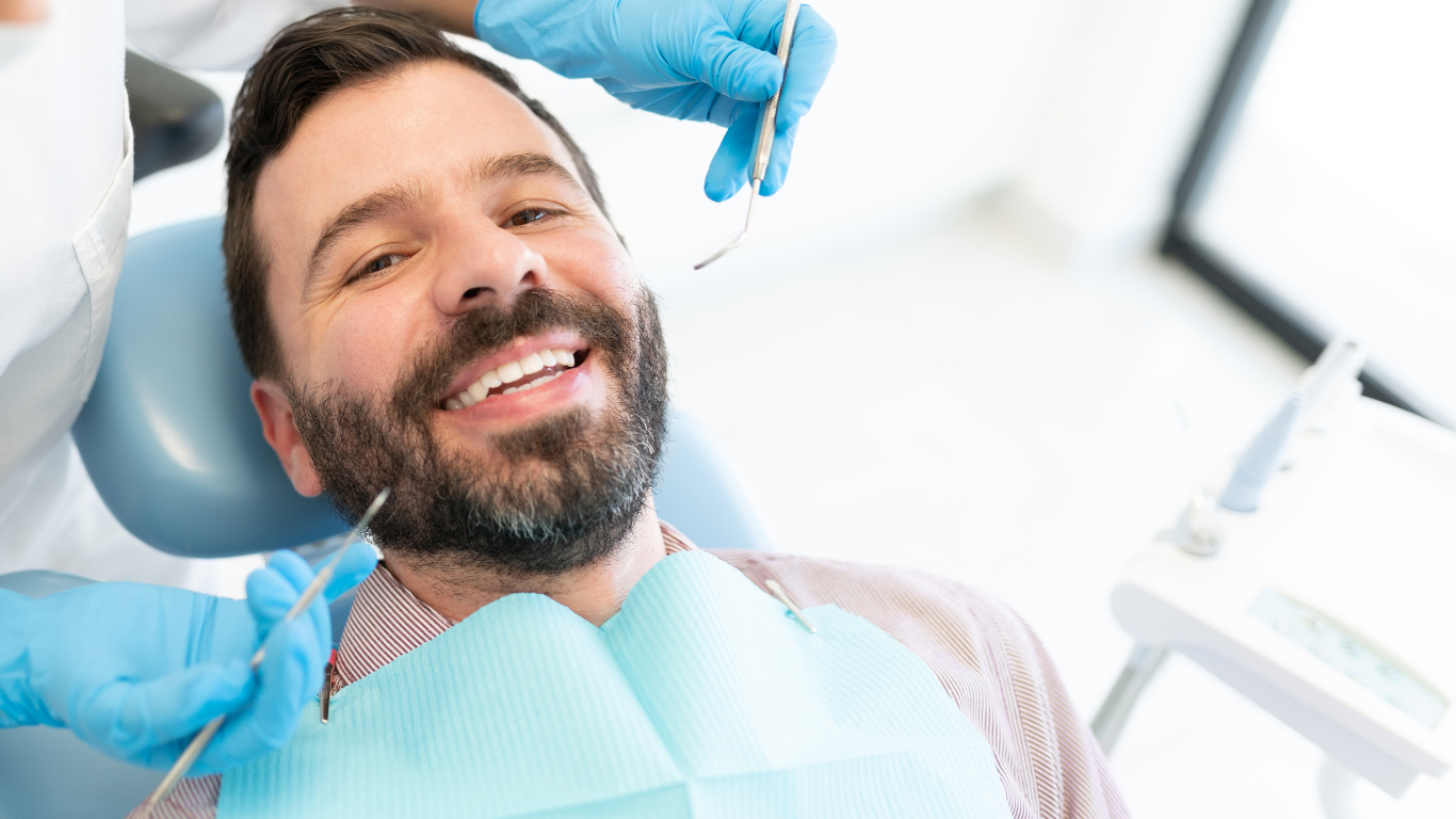 Man in dentist chair, smiling, dental tools near his mouth.