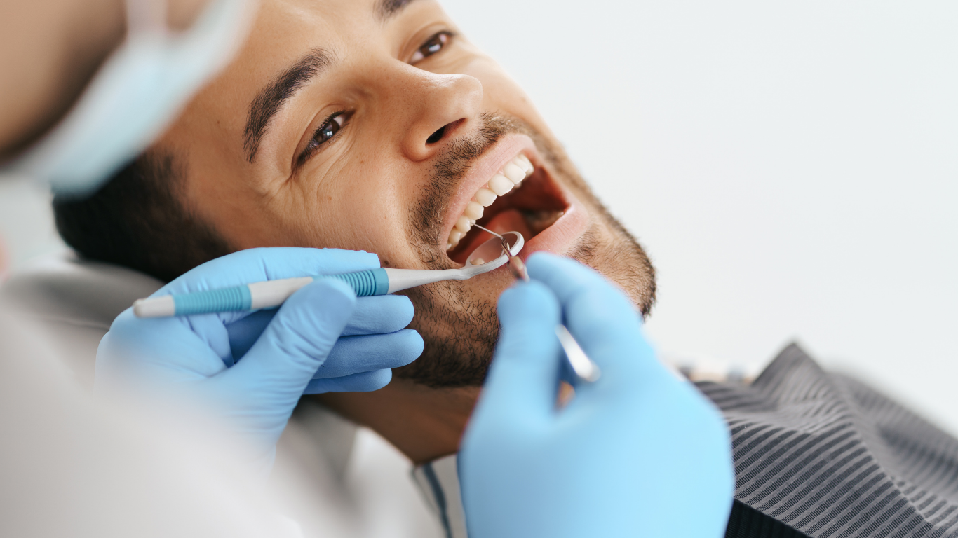 Man at dentist, mouth open, being examined with tools by gloved hands.