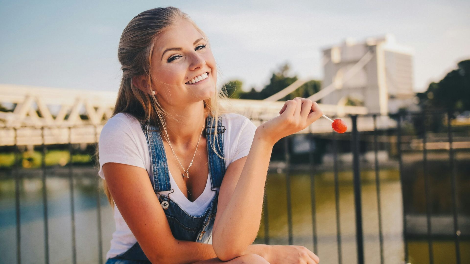 Woman with blonde hair smiles, holding a lollipop. She wears a white shirt and denim overalls, seated outdoors near water and a bridge.