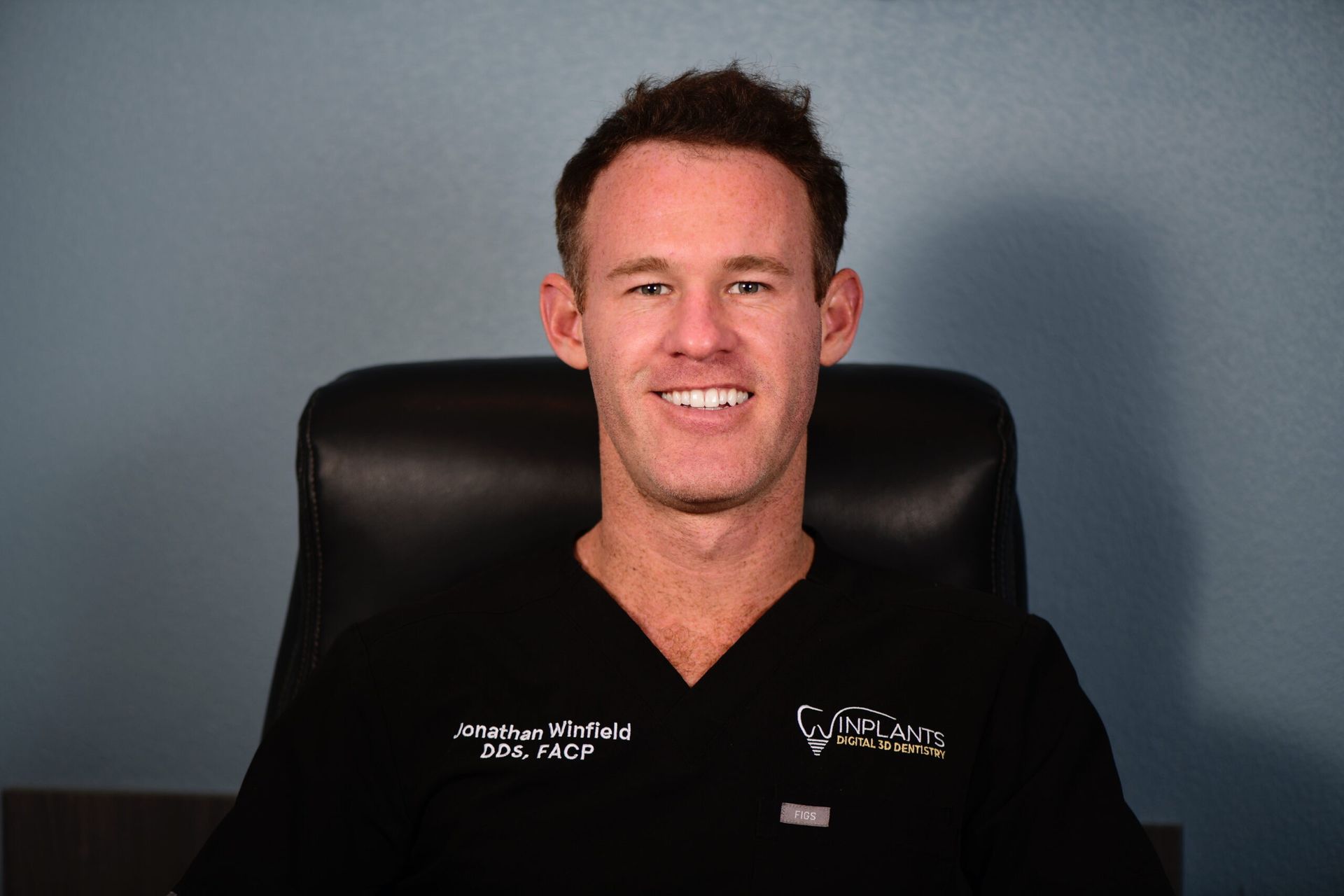 Man in black scrubs, smiling, seated in a chair, with a logo on the scrubs. Blue background.