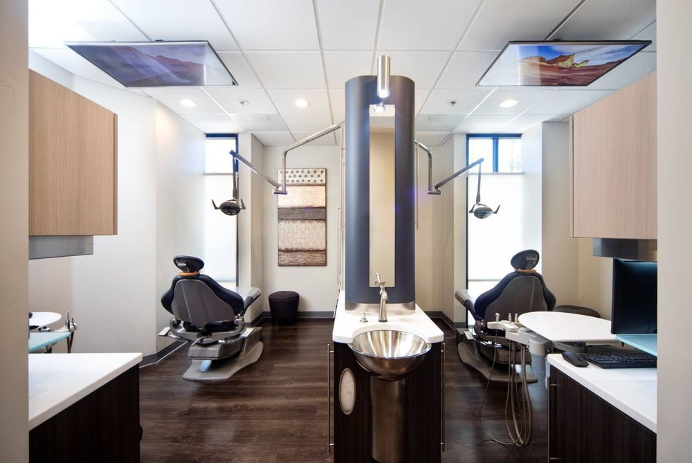 Dental operatory with two chairs, overhead lights, sinks, cabinetry, and TVs.