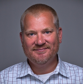 A man with a beard is wearing a plaid shirt and smiling for the camera.