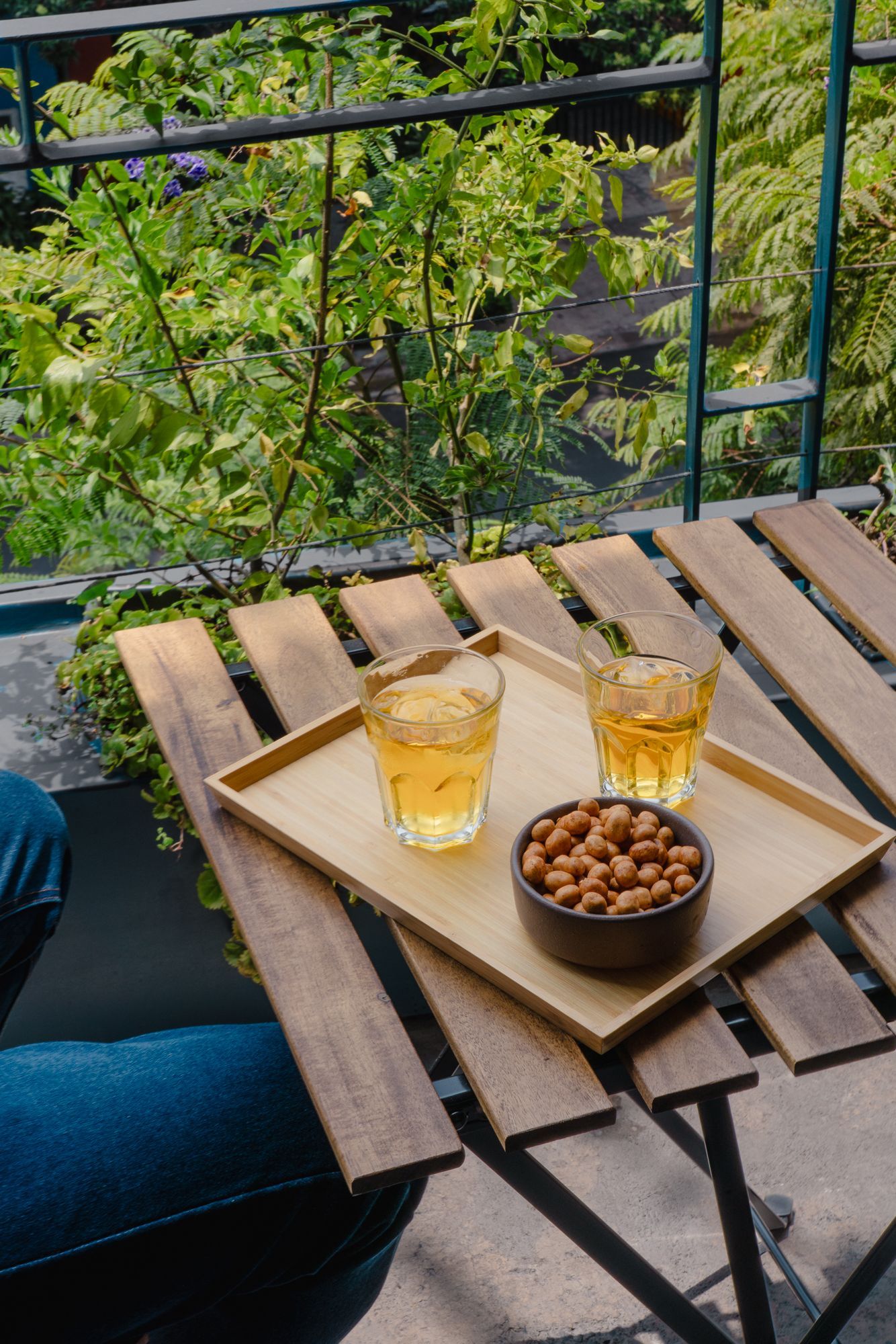 Two drinks and peanuts on a wooden tray atop a small table on a balcony overlooking lush greenery.
