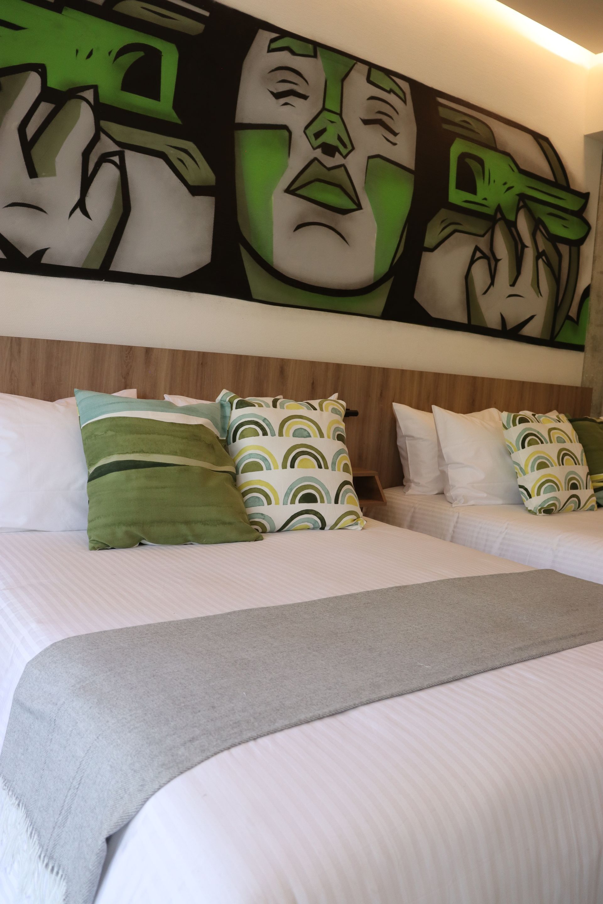 Two beds with white linens, gray runner, decorative pillows, and a mural of a face holding green guns.