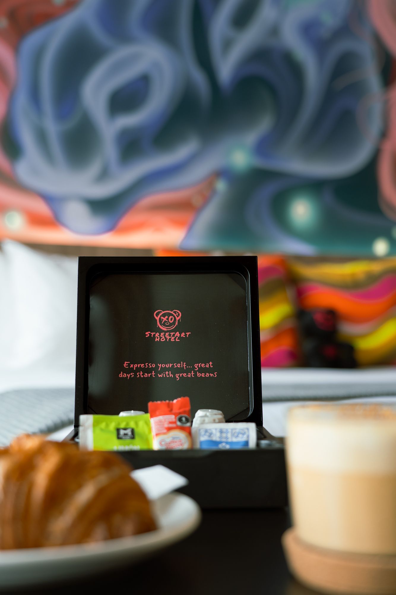 Breakfast tray with croissant, tea, and coffee in a hotel room with colorful artwork in the background.