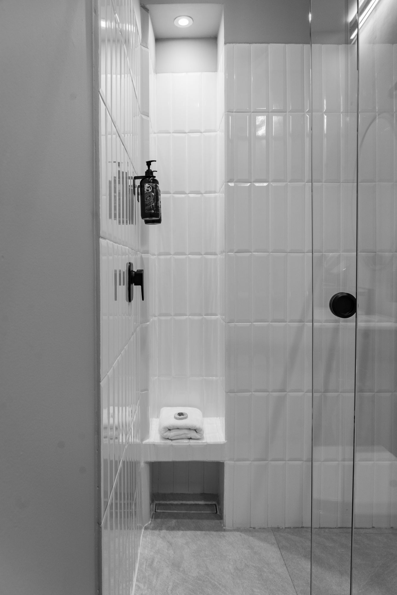 A monochrome shower with white tiles, a built-in shelf, glass door, and black fixtures.