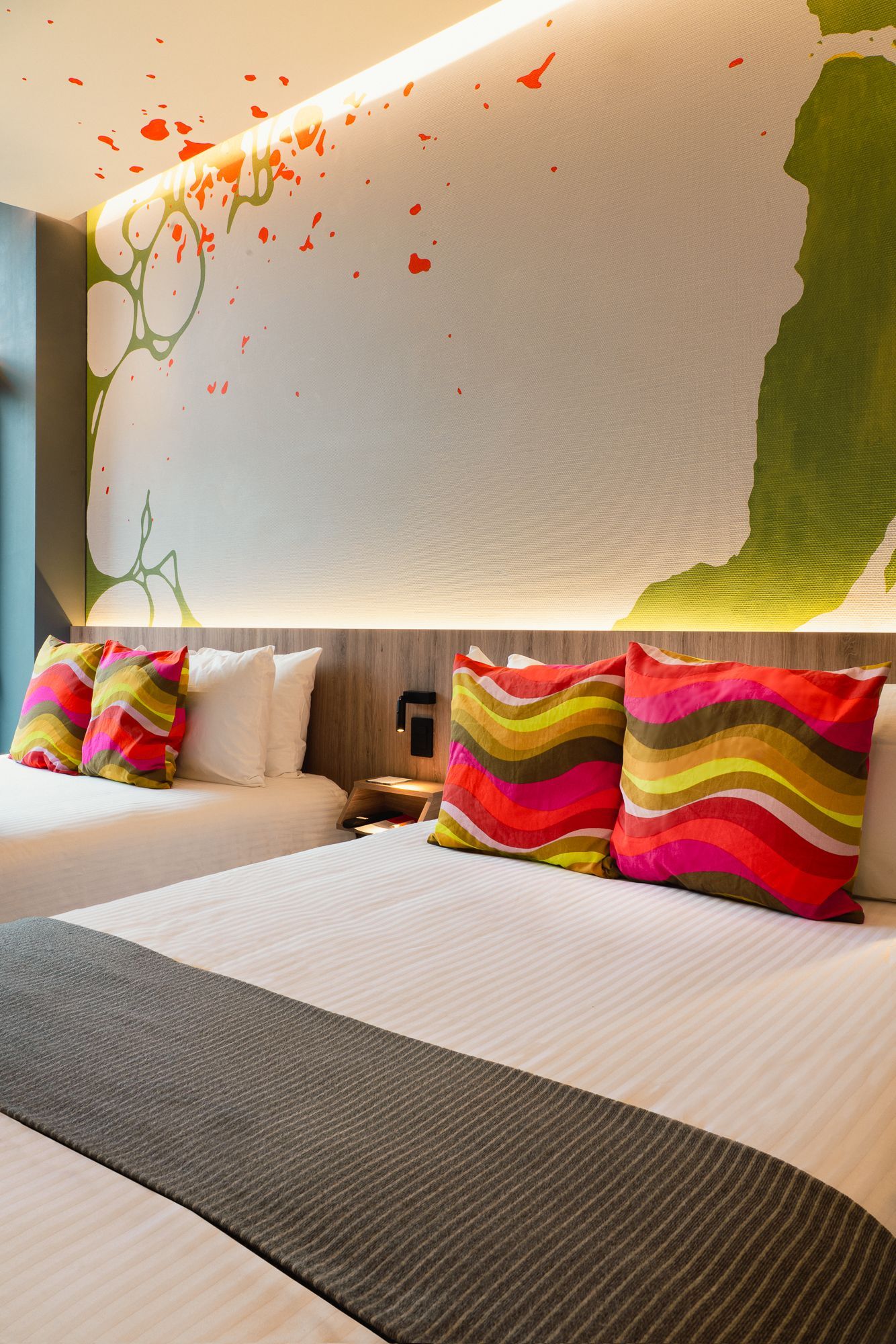 Two beds with decorative pillows against a wall mural with orange and green paint splatters.