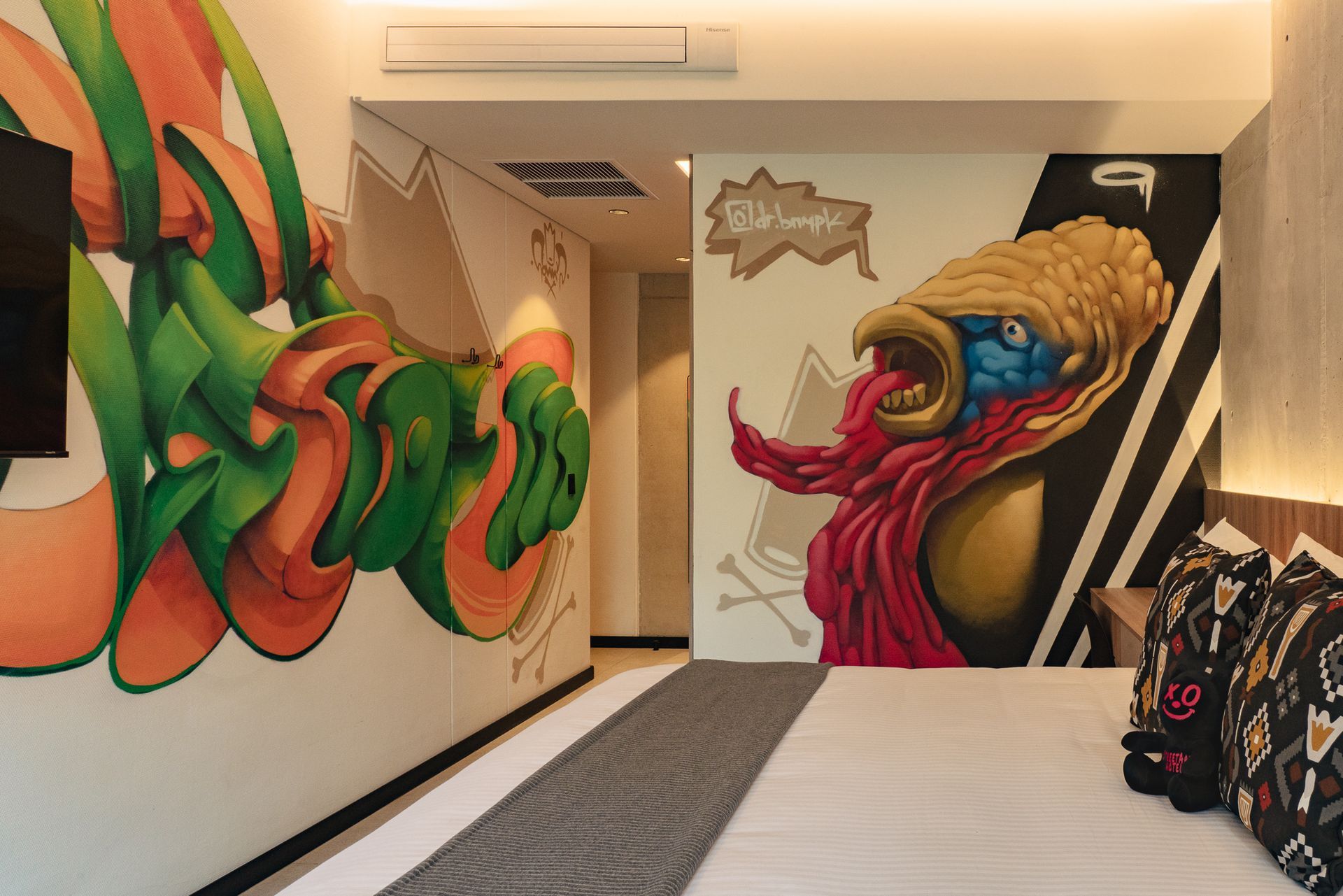 Hotel room with graffiti art on the walls; a bed with patterned pillows is in the foreground.