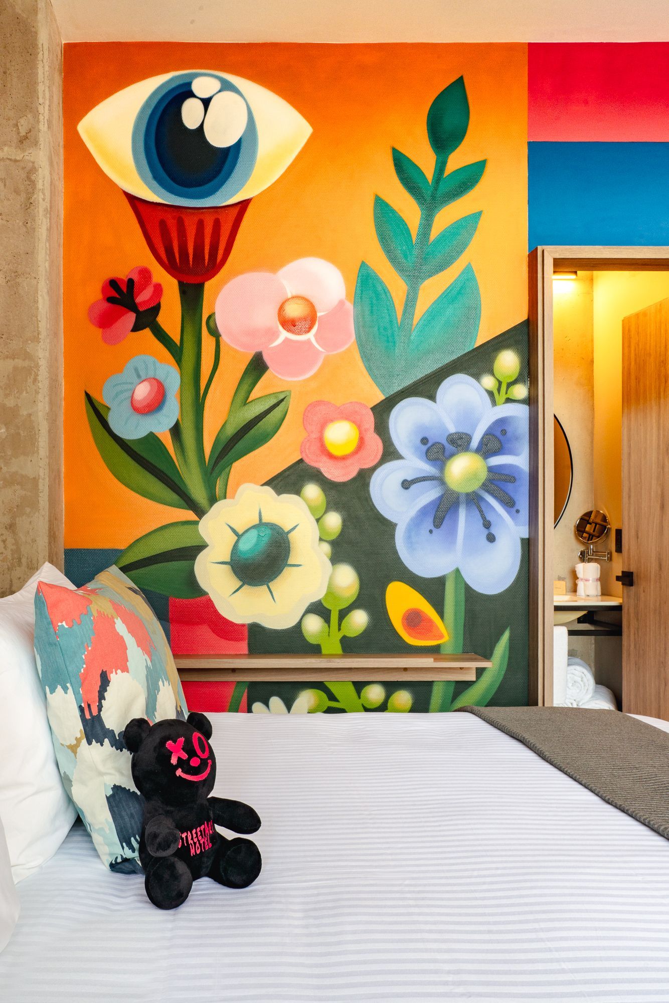 Bedroom with mural of a stylized eye and flowers; bed with pillows and teddy bear.