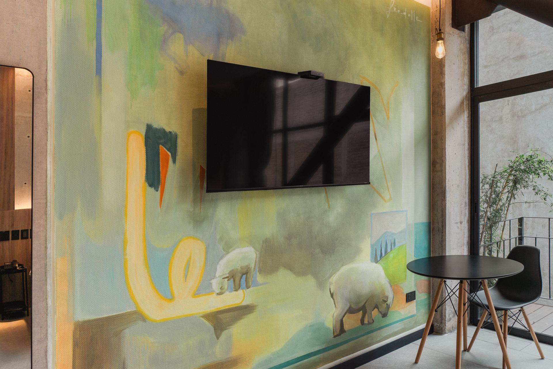 A TV mounted on a colorful mural with sheep, next to a black table and chair, by a window.