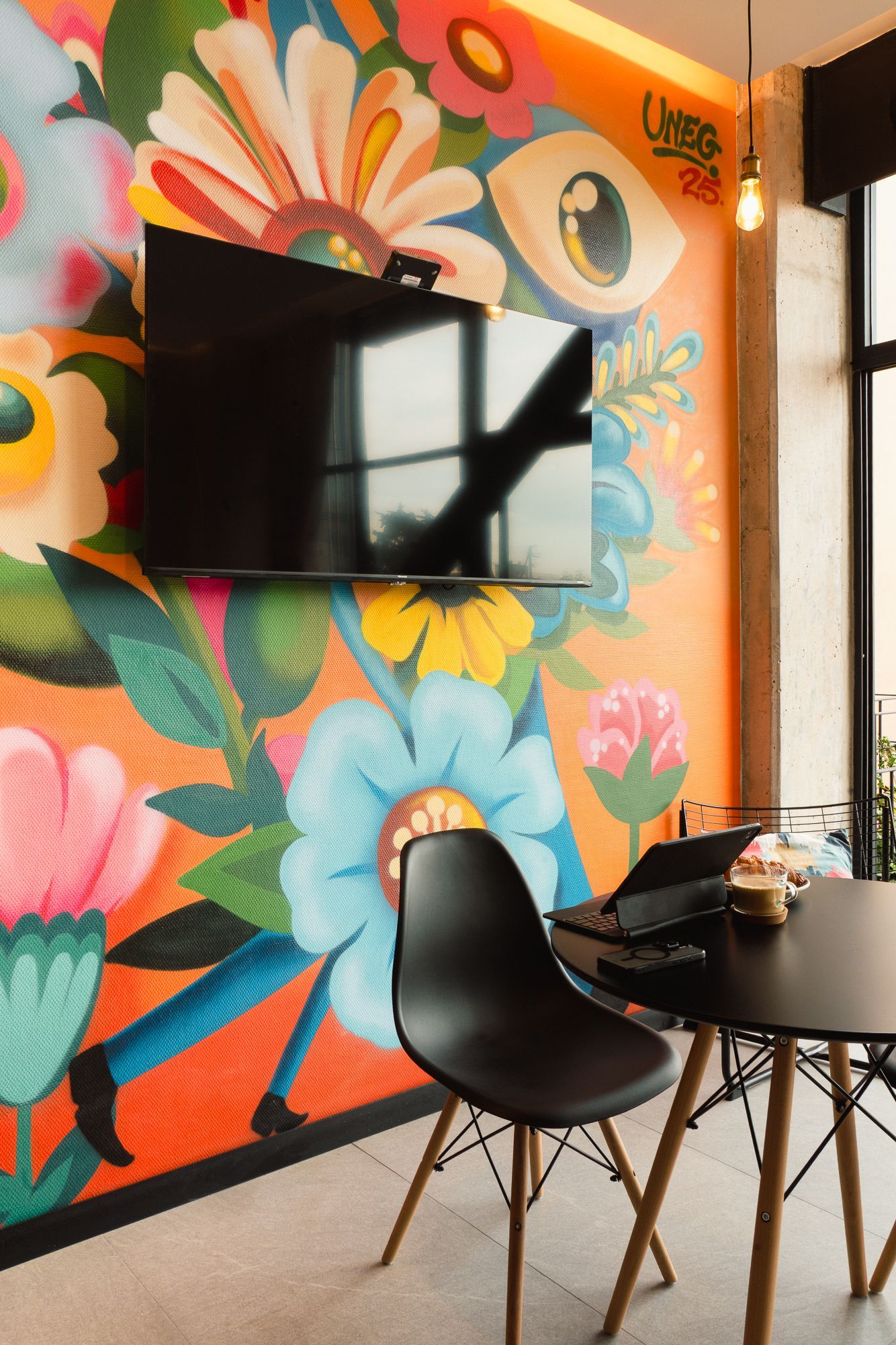 Black chair and table with laptop in front of a colorful flower mural with a mounted TV.