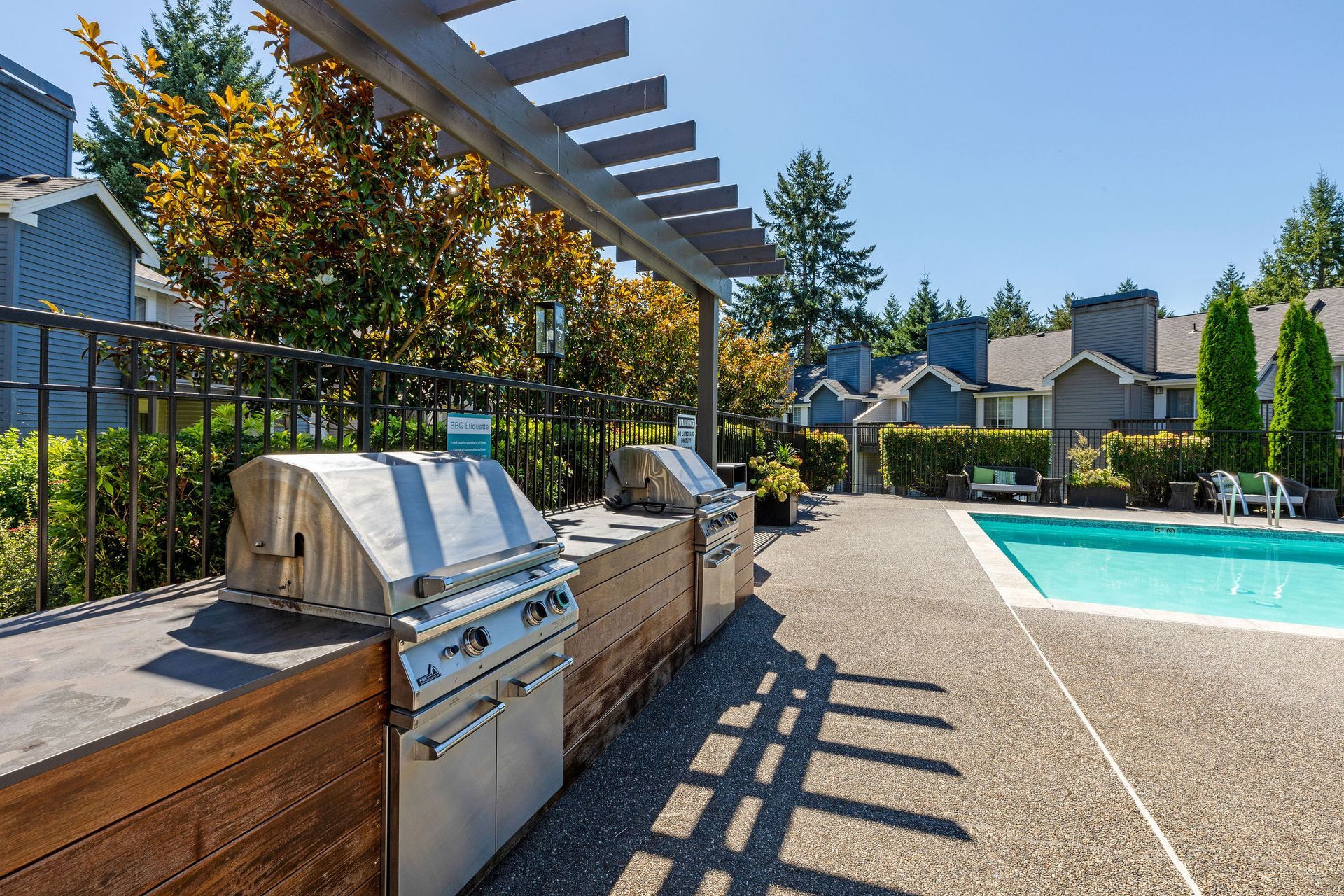 Grill area with stainless steel grills next to a pool. Wooden pergola and patio in the background.