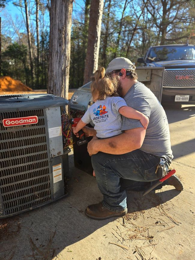 Man kneeling and hugging a small child beside a truck grille in a sunny wooded driveway.