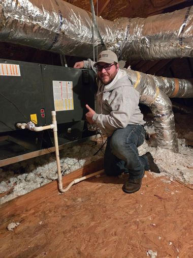 Worker kneeling beside HVAC unit in an unfinished attic, inspecting ductwork and piping.
