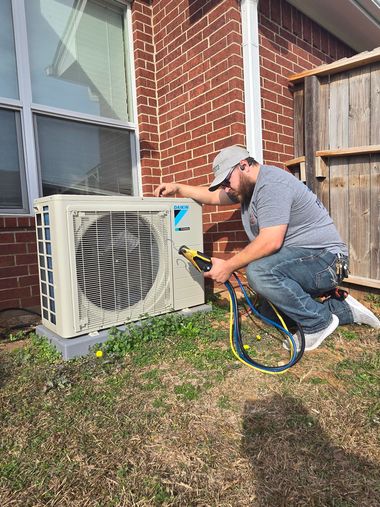 Technician kneeling by an outdoor HVAC unit, checking hoses beside a brick house