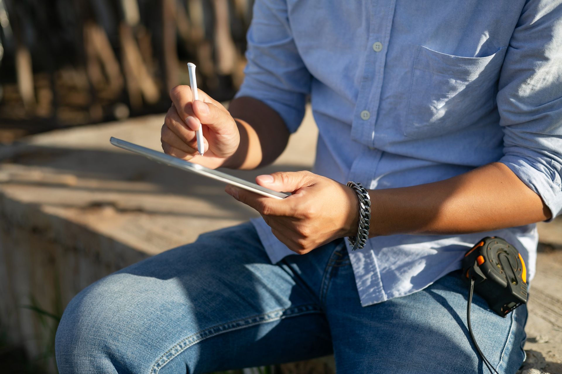 Person in blue shirt and jeans using a stylus on a tablet outdoors. A measuring tape is visible.