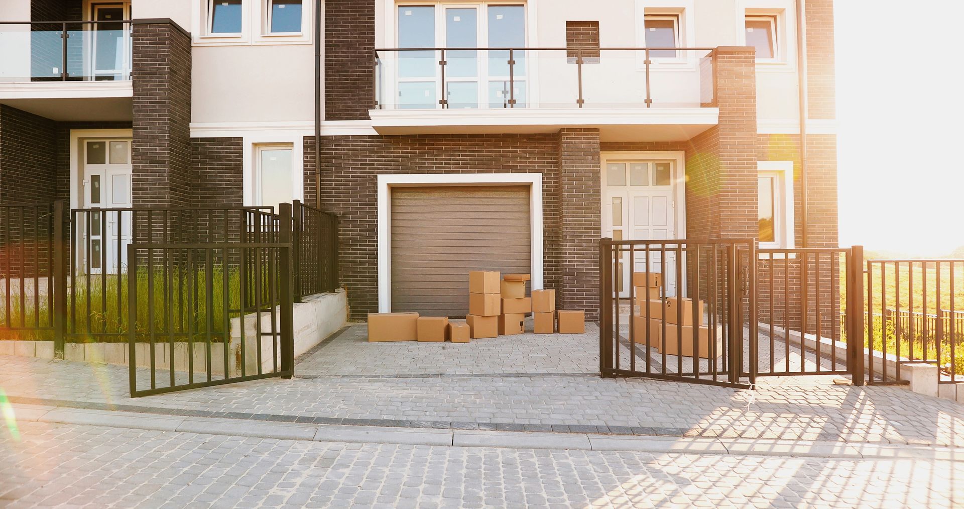 Brown boxes stacked outside a house with a closed garage door and open gate; sunlight.