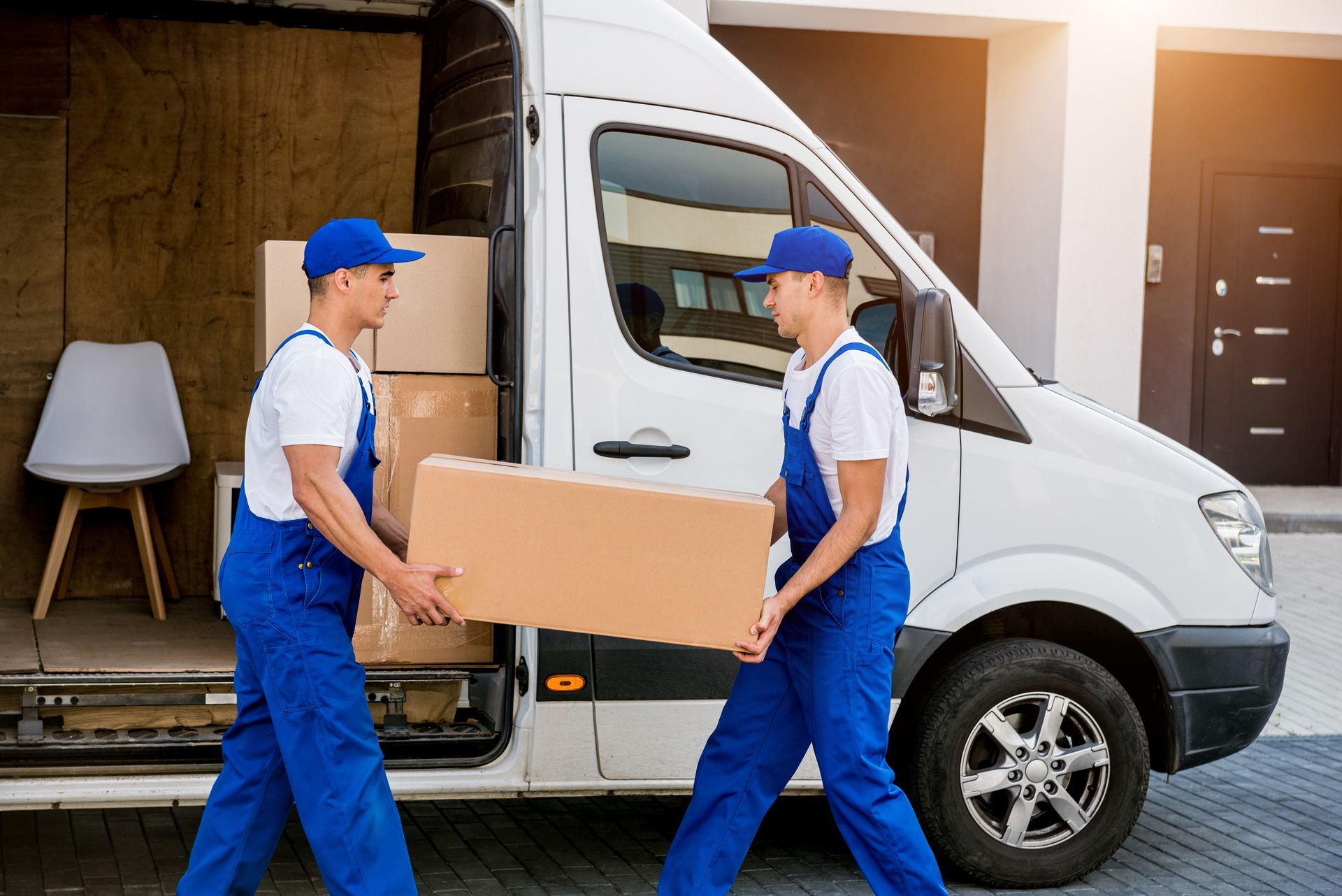 Two movers in blue coveralls and hats carry a large box to a white moving van.