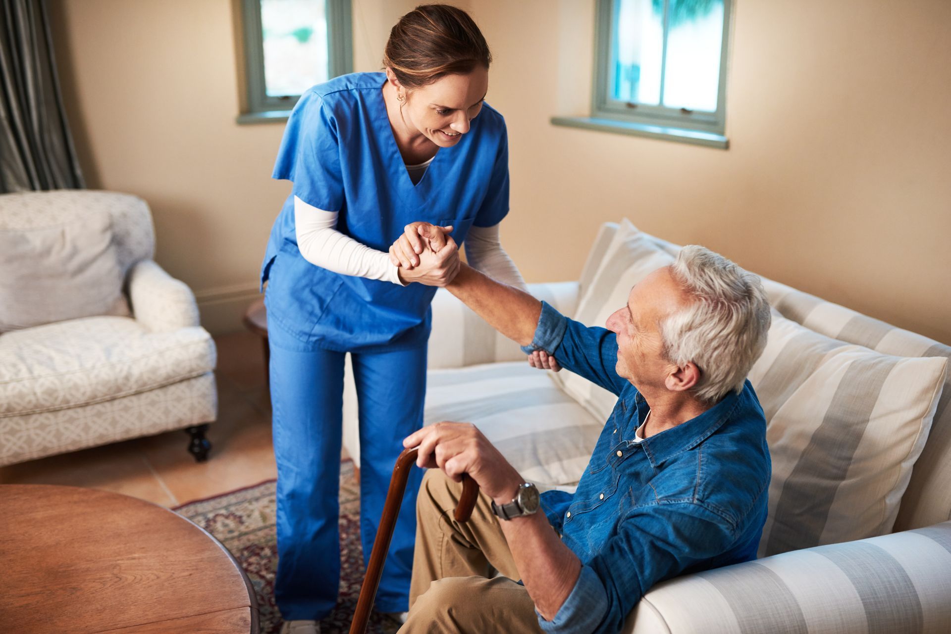 Nurse assists elderly person sitting on a sofa, helping him stand using a cane.