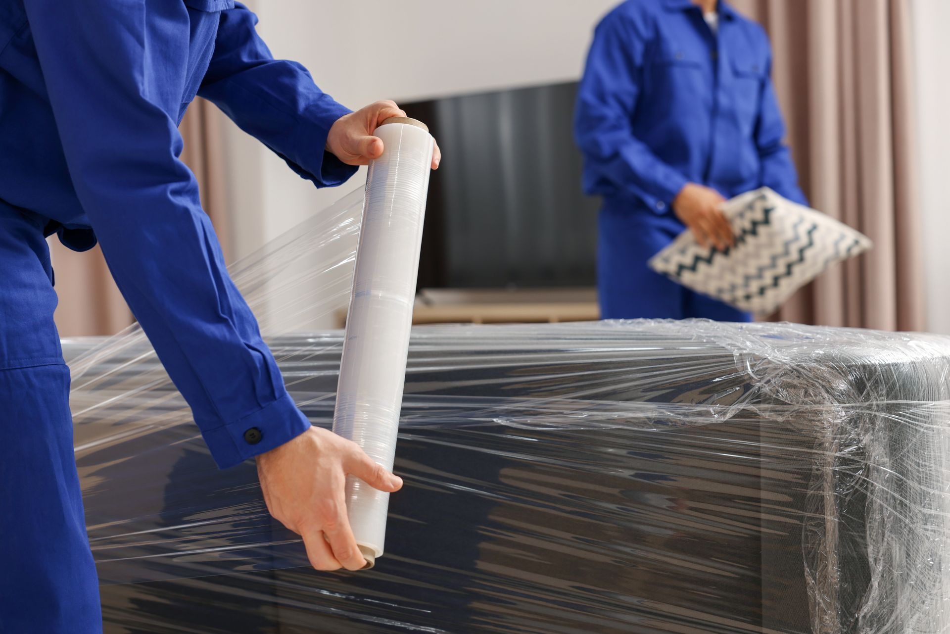 Movers wrapping furniture with clear plastic in a home; blue uniforms.